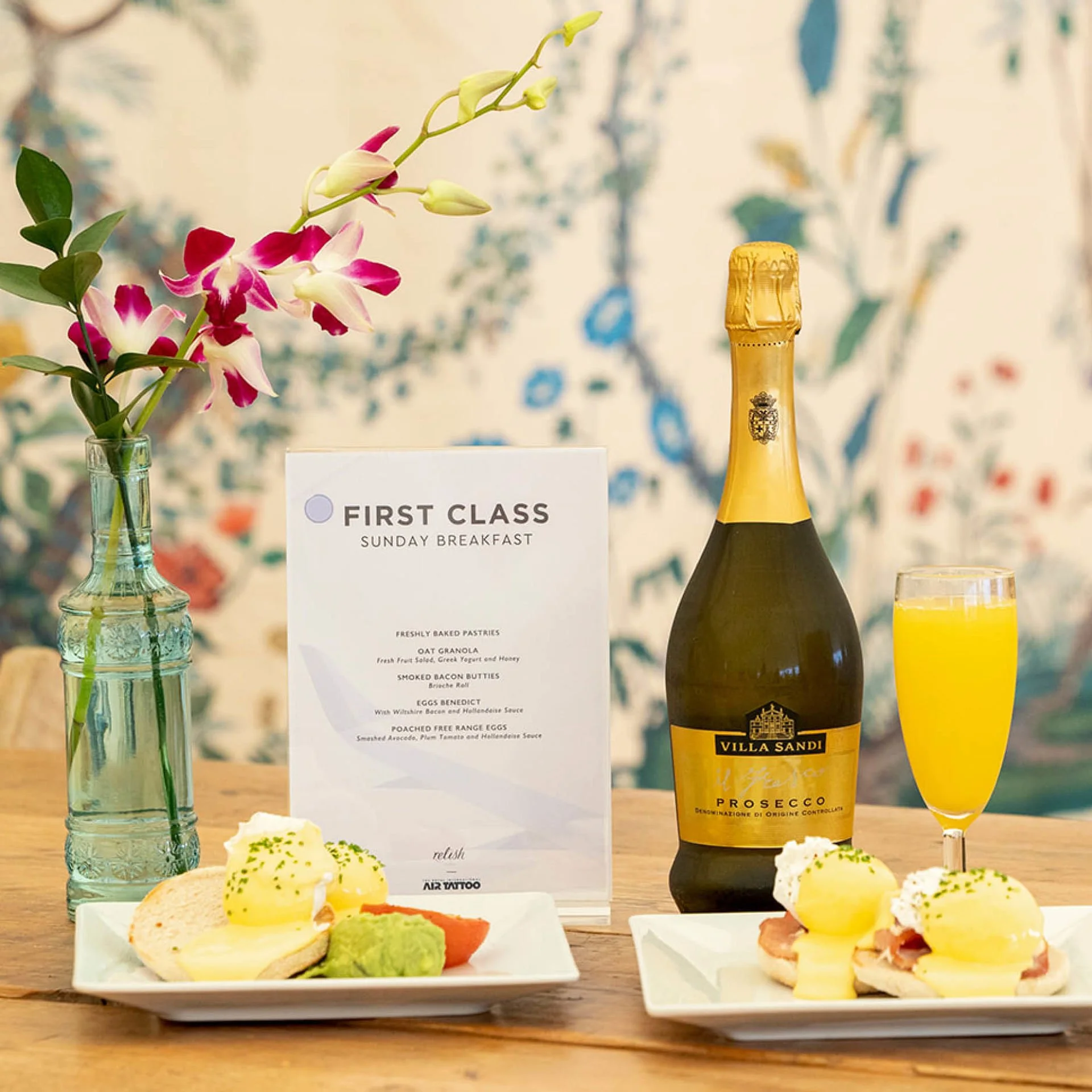 A breakfast table with a vase of pink and white flowers, a menu titled 'First Class Sunday Breakfast,' a bottle of Villa Sandi Prosecco, a glass of orange juice, and plates of eggs Benedict with bacon and toast, garnished with parsley.
