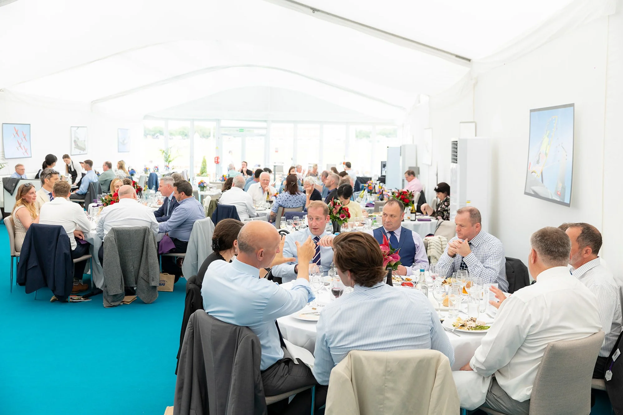 People attending a banquet in a bright white tent, sitting at round tables decorated with floral centerpieces, having conversations and enjoying a meal.