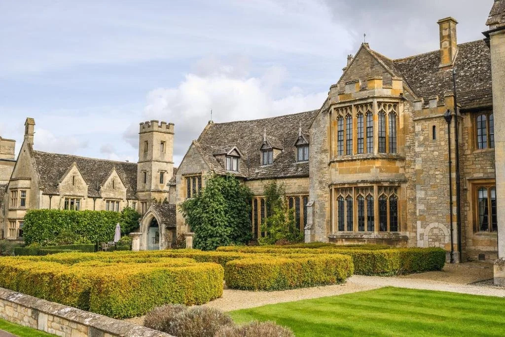 A historic European castle with stone walls and large windows, surrounded by well-maintained gardens and hedges.