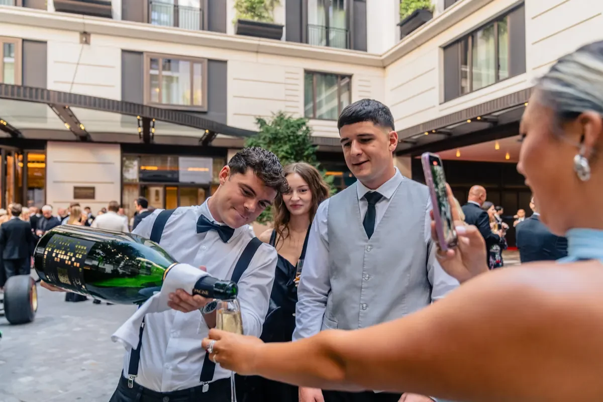 A group of people dressed in formal attire at an outdoor event, with a woman pouring champagne into a glass held by a young man, as others look on and smile.