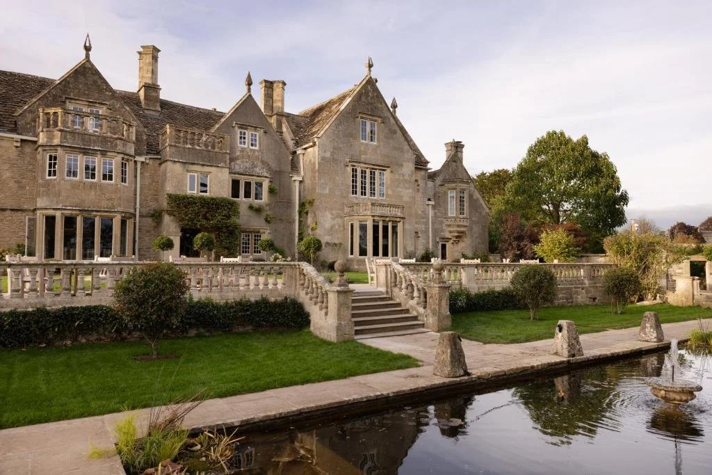 A historic stone mansion with multiple gabled roofs, surrounded by a landscaped garden with trees and shrubs, a stone staircase leading to a terrace, and a small pond with a fountain in the foreground.