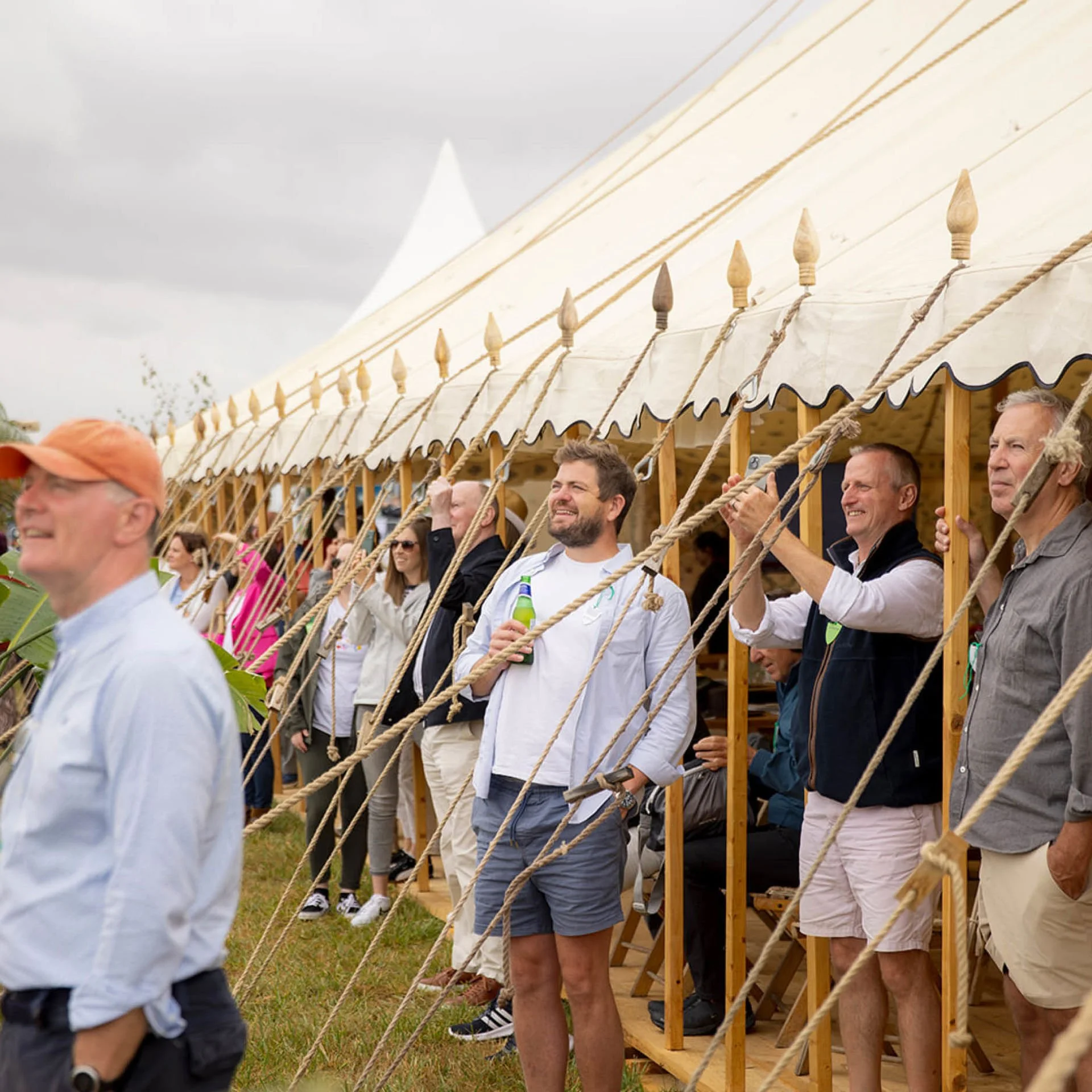 People standing outside a large tent at an outdoor event, some holding drinks, all smiling and enjoying the gathering.