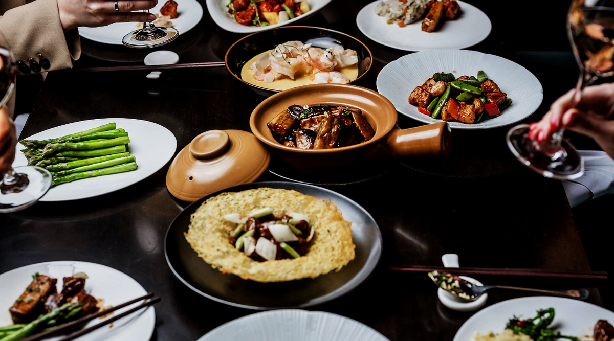 A table with various Asian dishes, including grilled asparagus, a rice and vegetable dish, a fried egg dish, a bowl of braised meat, a seafood dish in a bowl, and multiple plates of stir-fry, with people holding wine glasses.