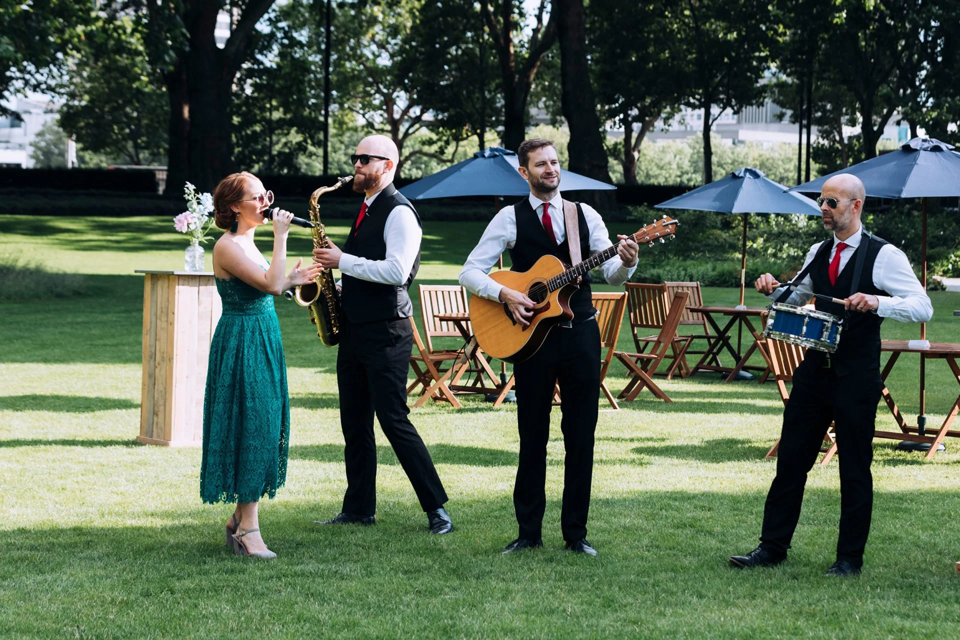 A band performing outdoors on a lush green lawn, consisting of a woman singing into a microphone, a man with a saxophone, a man playing an acoustic guitar, and a man with a snare drum. There are empty chairs and tables with umbrellas in the background, surrounded by trees.