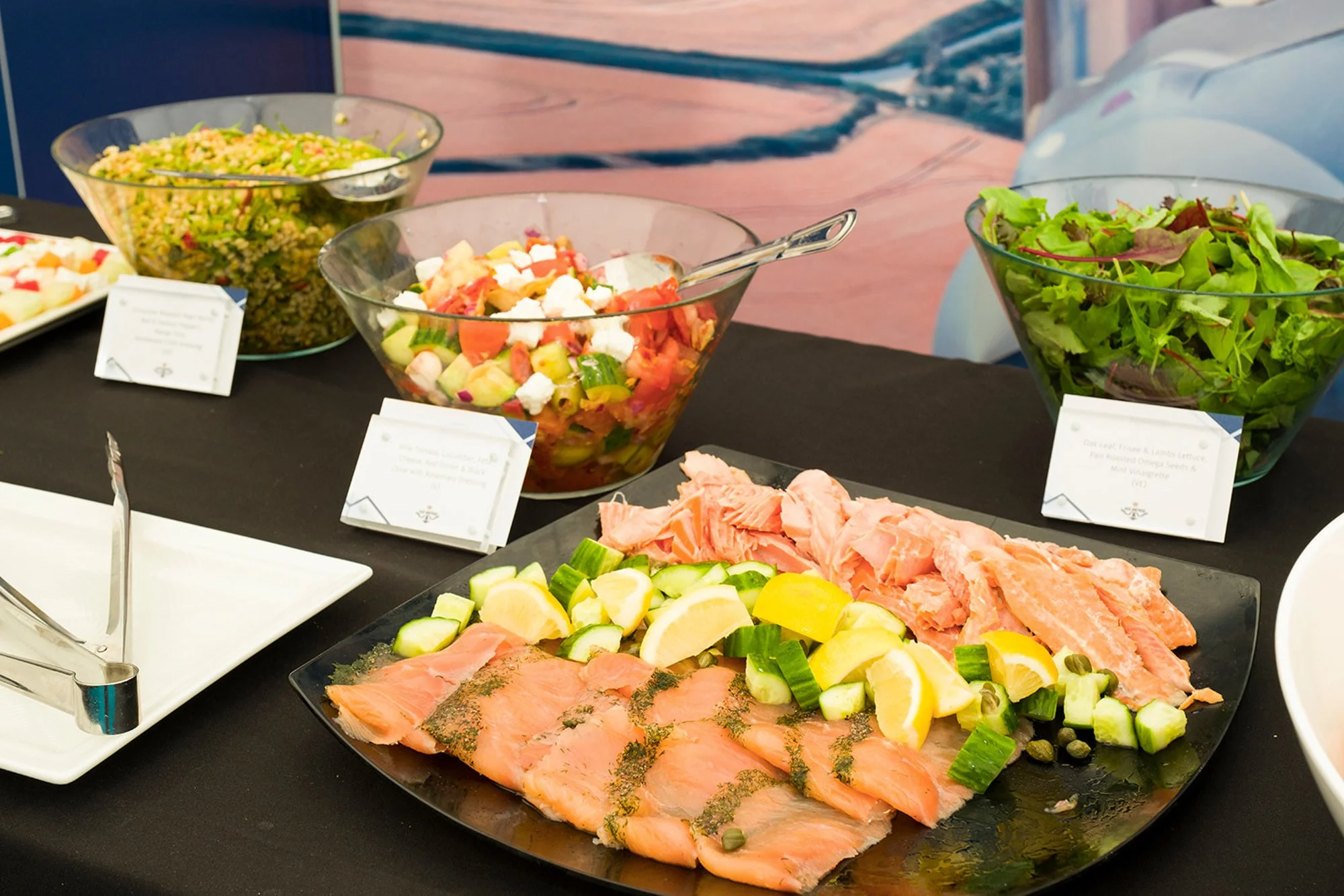 A buffet table with slices of smoked salmon garnished with lemon wedges and cucumbers, and three bowls of salad containing mixed greens, a Mediterranean-style salad with tomatoes and feta, and a grain salad with herbs.