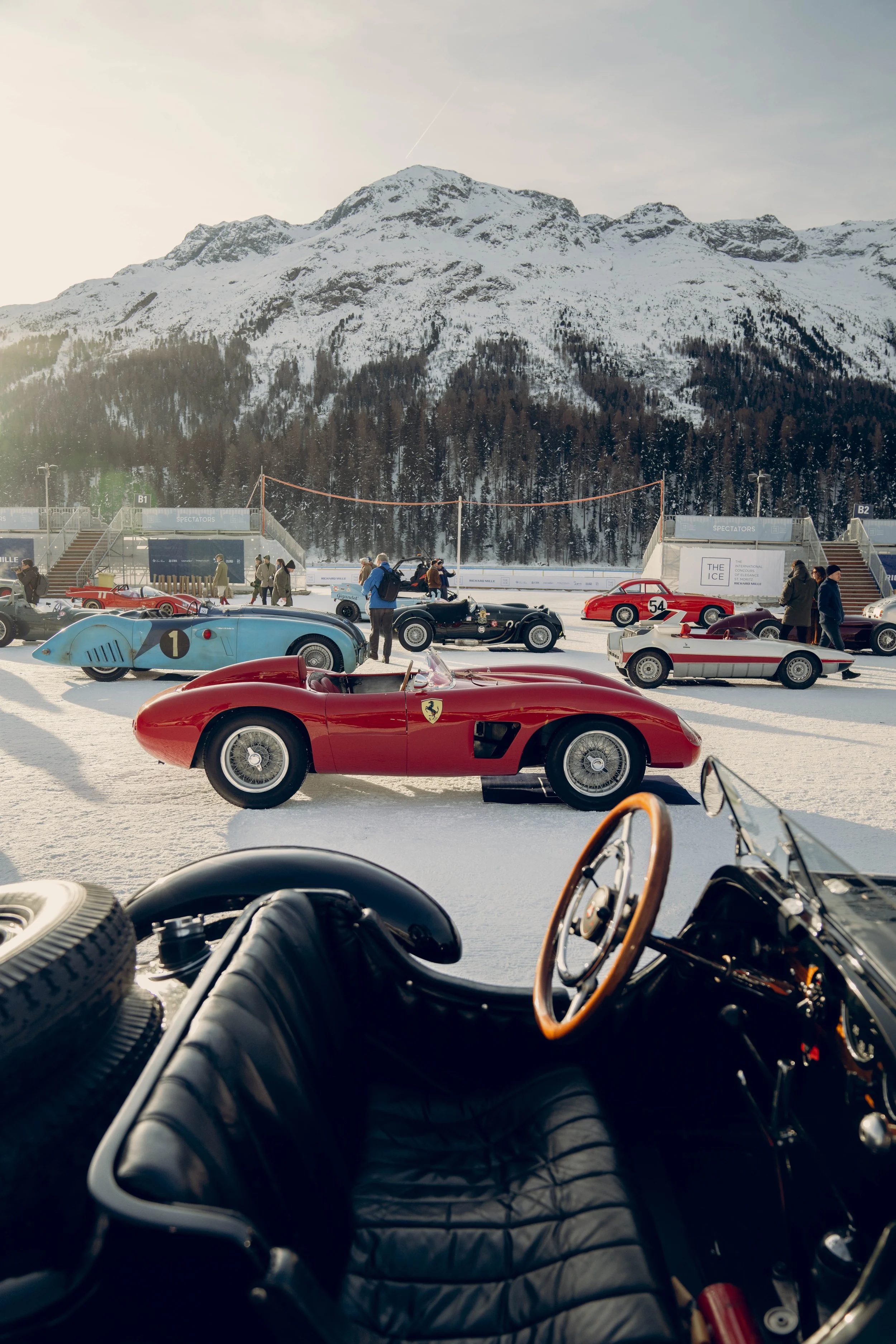 A classic car exhibition on snow with vintage racing cars, including a red Ferrari, set against snowy mountains and a clear sky.