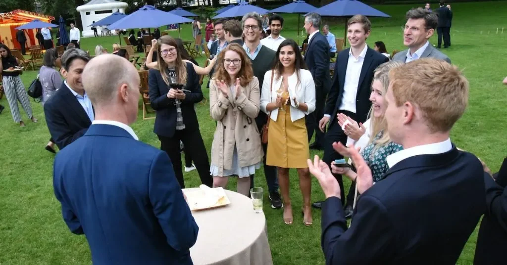 Group of people in formal attire gathered outdoors at a celebration or social event with a man at a table with a cake, others are smiling and clapping, some holding drinks, in a grassy area with umbrellas and additional tents.