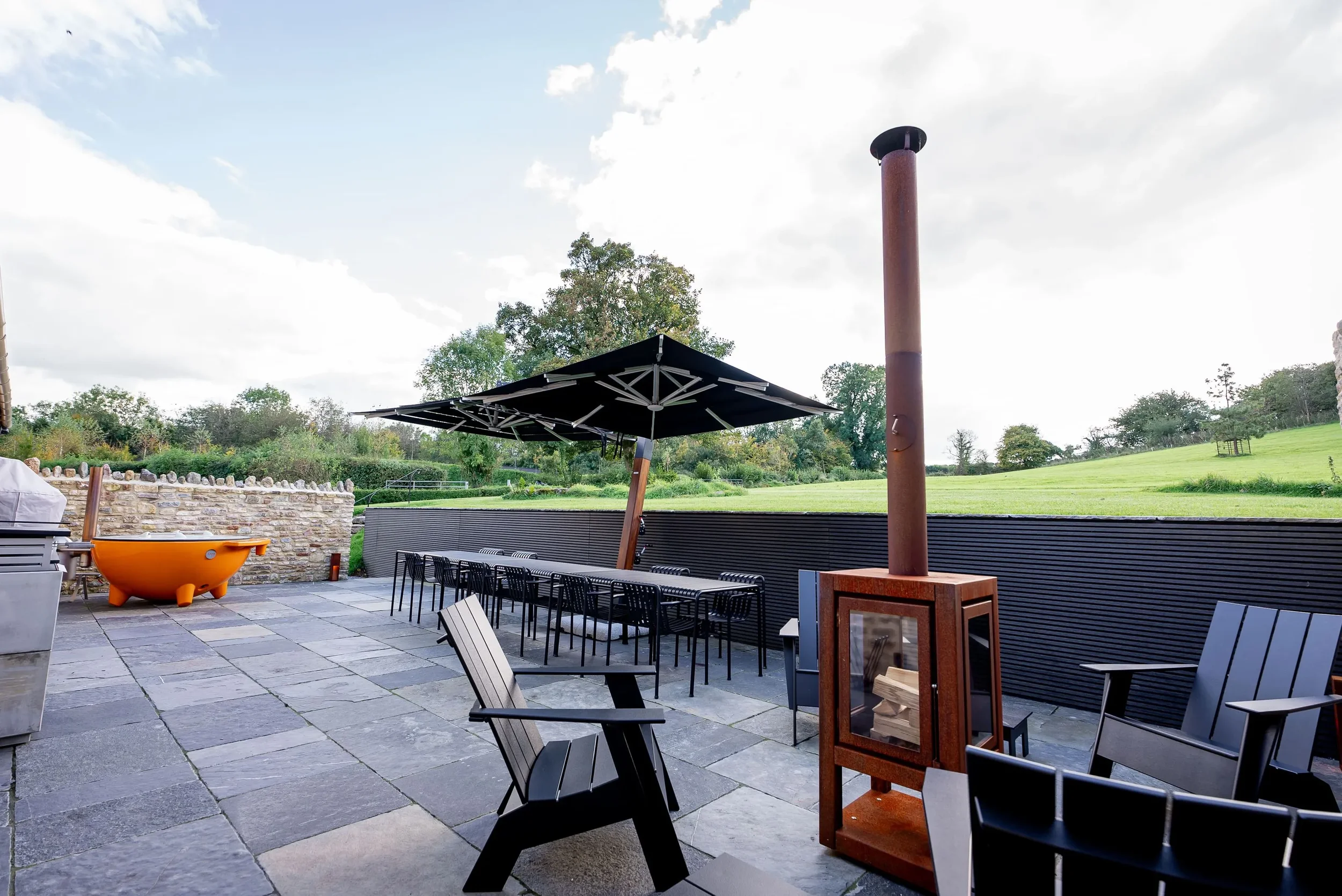 Outdoor patio with black Adirondack chairs, a wood-burning stove, large umbrella, a small orange bathtub, and a black fence overlooking a green field and trees under a blue sky.