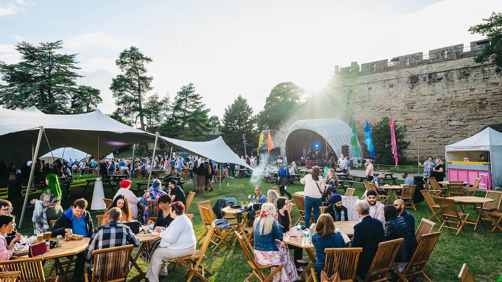 People gathering at an outdoor festival with tents, a stage, and picnic tables, in front of a large stone wall and trees, during sunset.