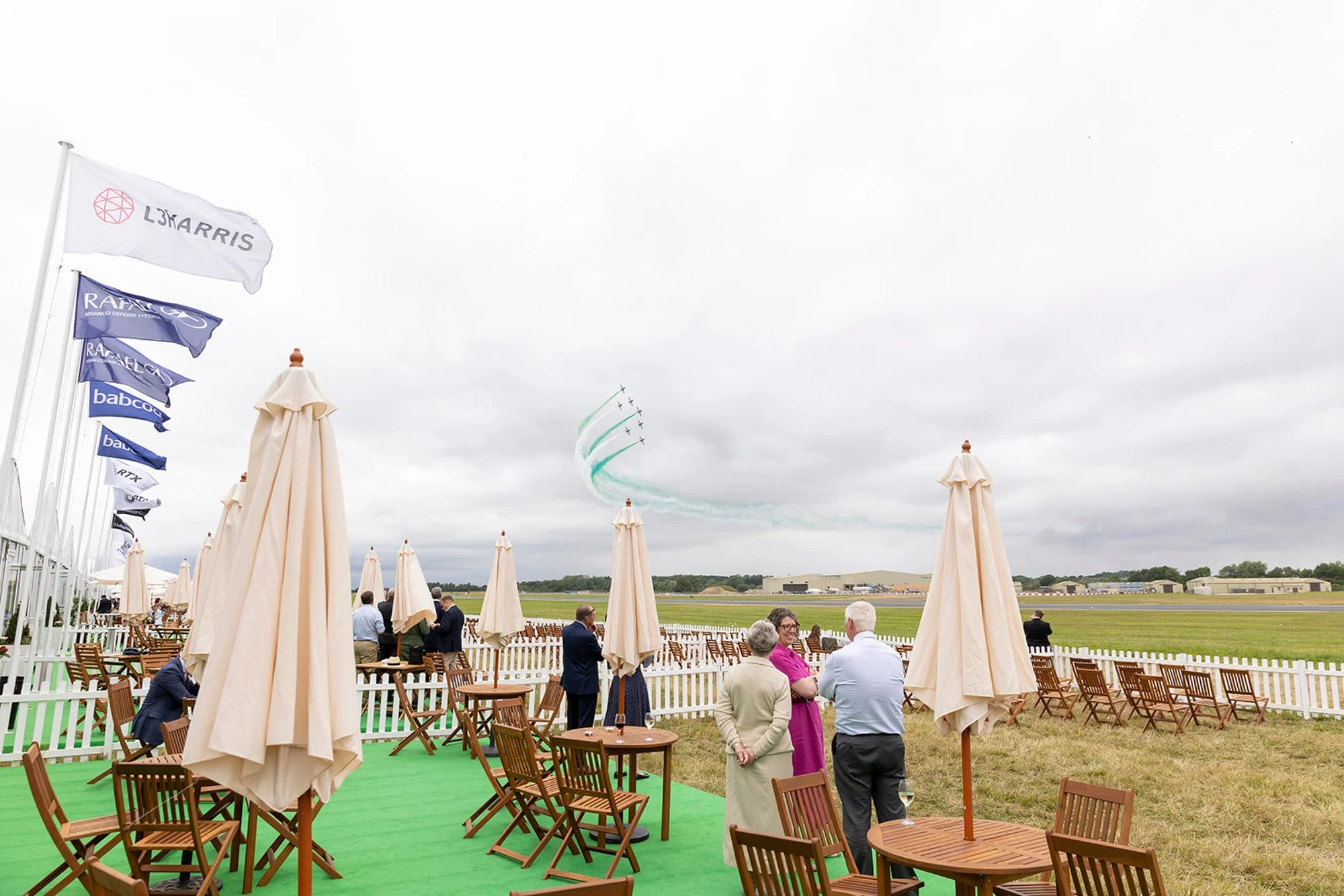 An outdoor event at an airfield with people standing and sitting by tables with beige umbrellas. There are flags on the left and aircraft flying in coordinated formation with green smoke trails in the sky.