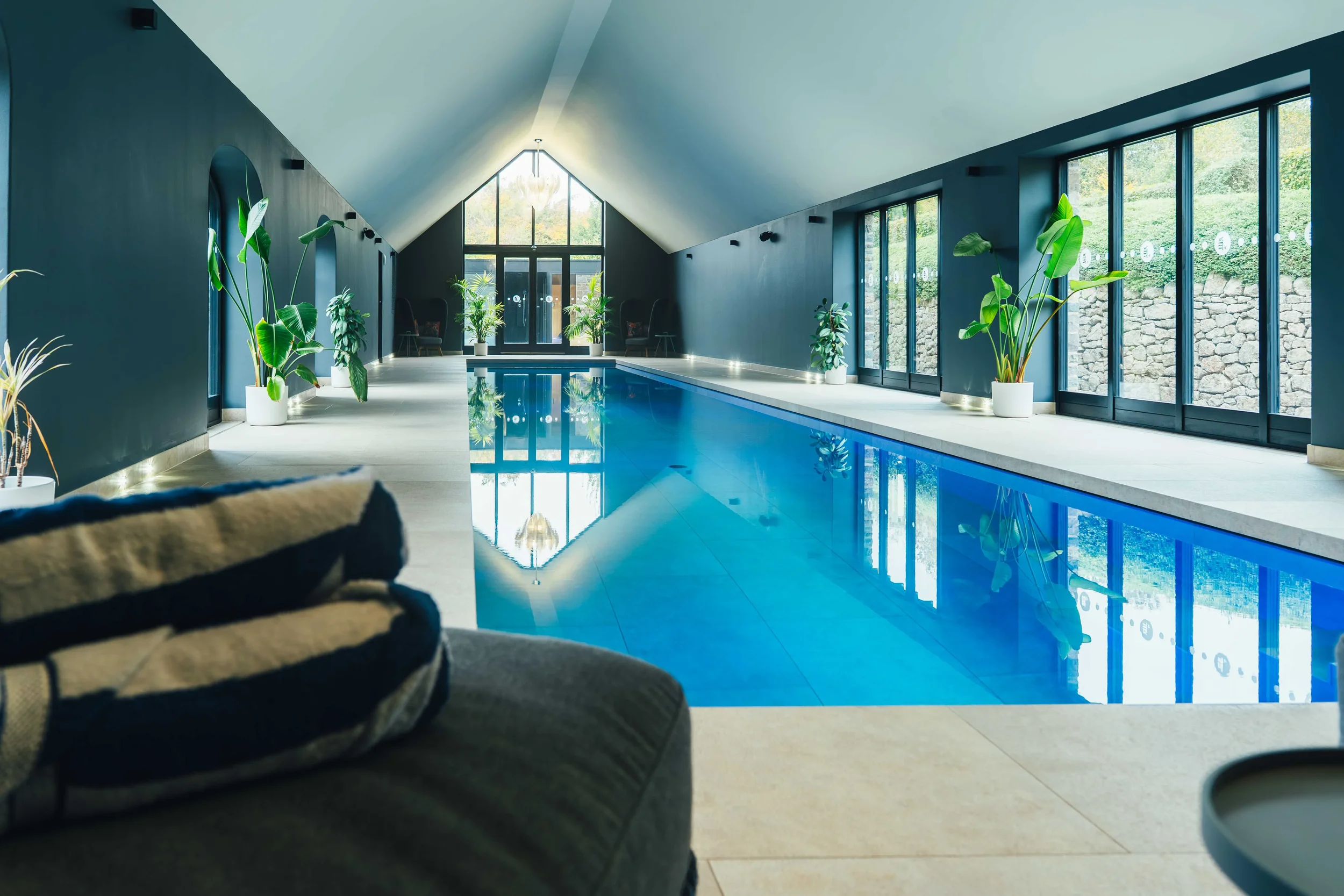 Indoor swimming pool with large windows, potted plants along the sides, and a high, sloped ceiling.