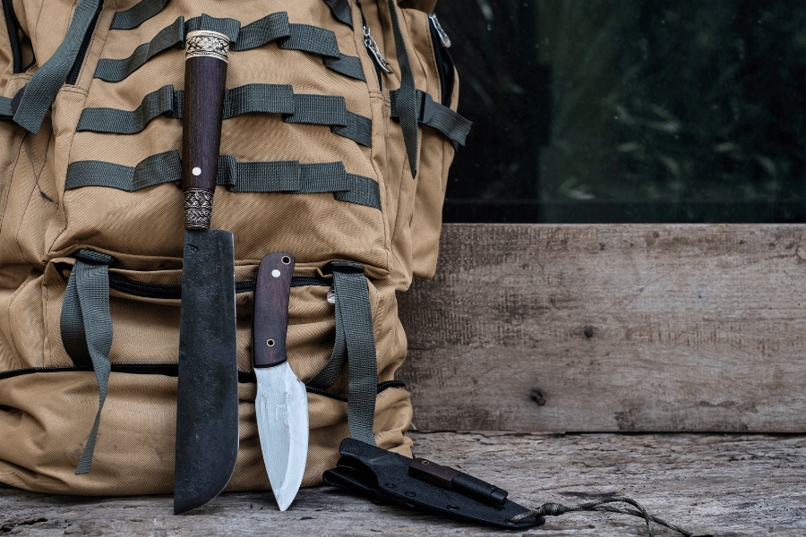 A backpack with two knives and a small axe attached, leaning against a wooden surface.