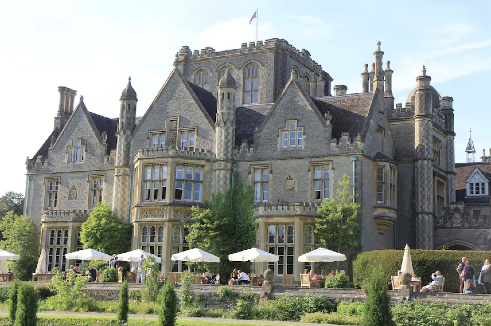 A large historic castle-like building made of stone with ornate windows and turrets. It has a castle feel with greenery and trees in the foreground and outdoor seating with white umbrellas where people are dining.