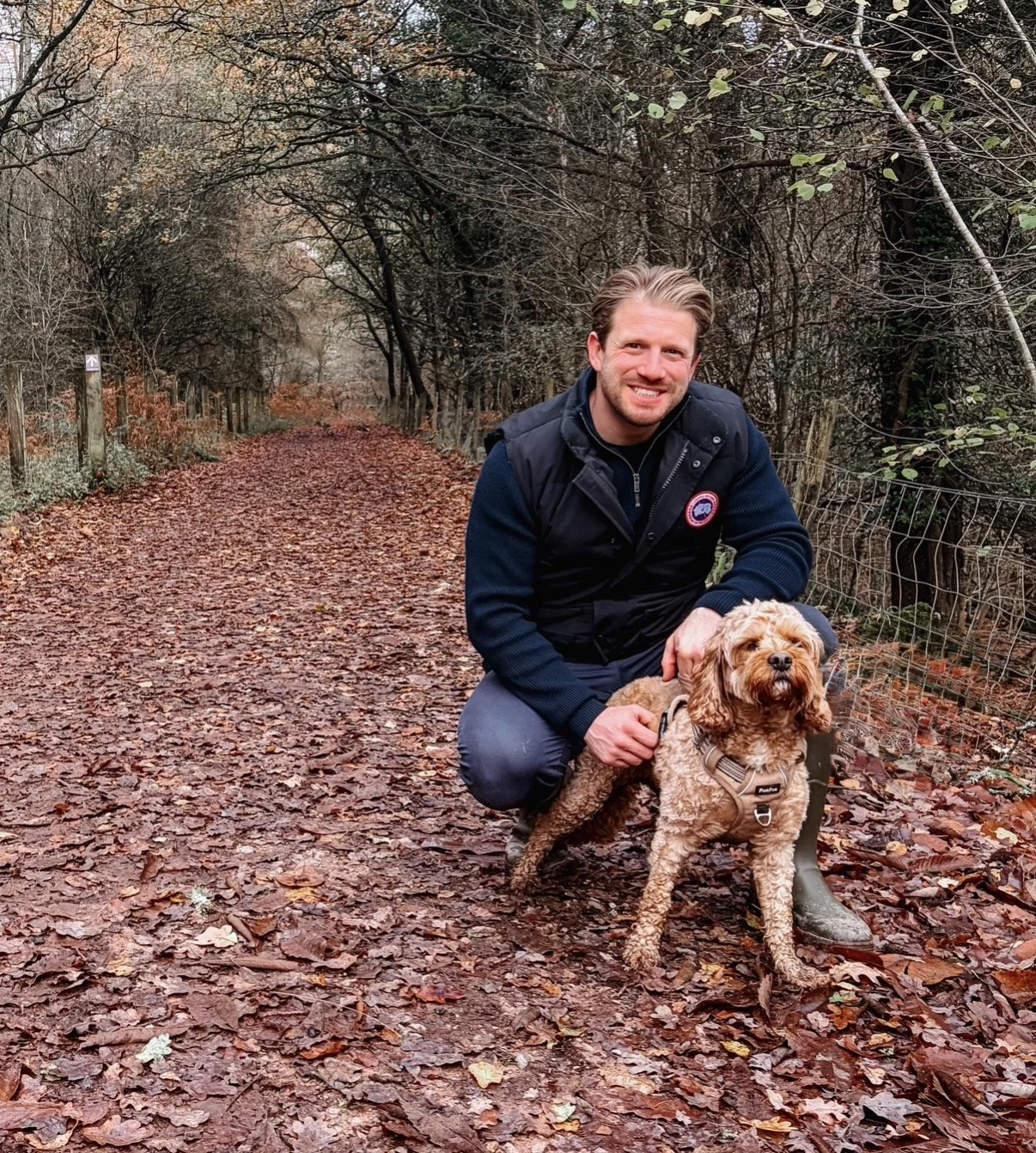 A man is kneeling on a leaf-covered forest trail next to a curly-haired dog wearing a harness.