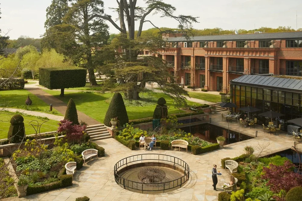 A landscaped patio with benches, a small round fountain, and lush greenery including topiary, trees, and garden beds. In the background, there's a red brick building and a glass-covered seating area.