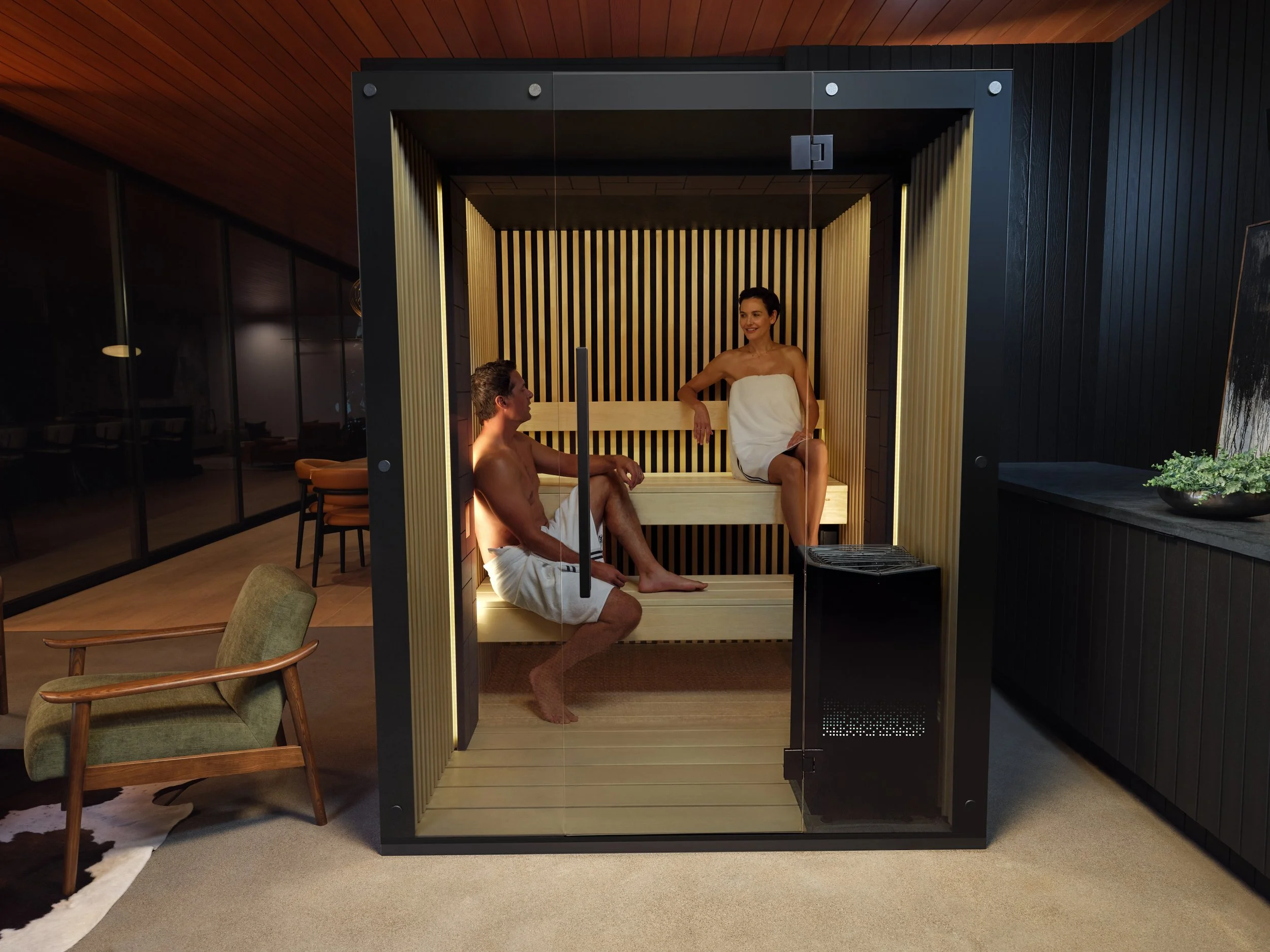 A man and woman in a modern sauna room with wooden interiors, sitting on the benches wrapped in towels, engaged in a conversation.