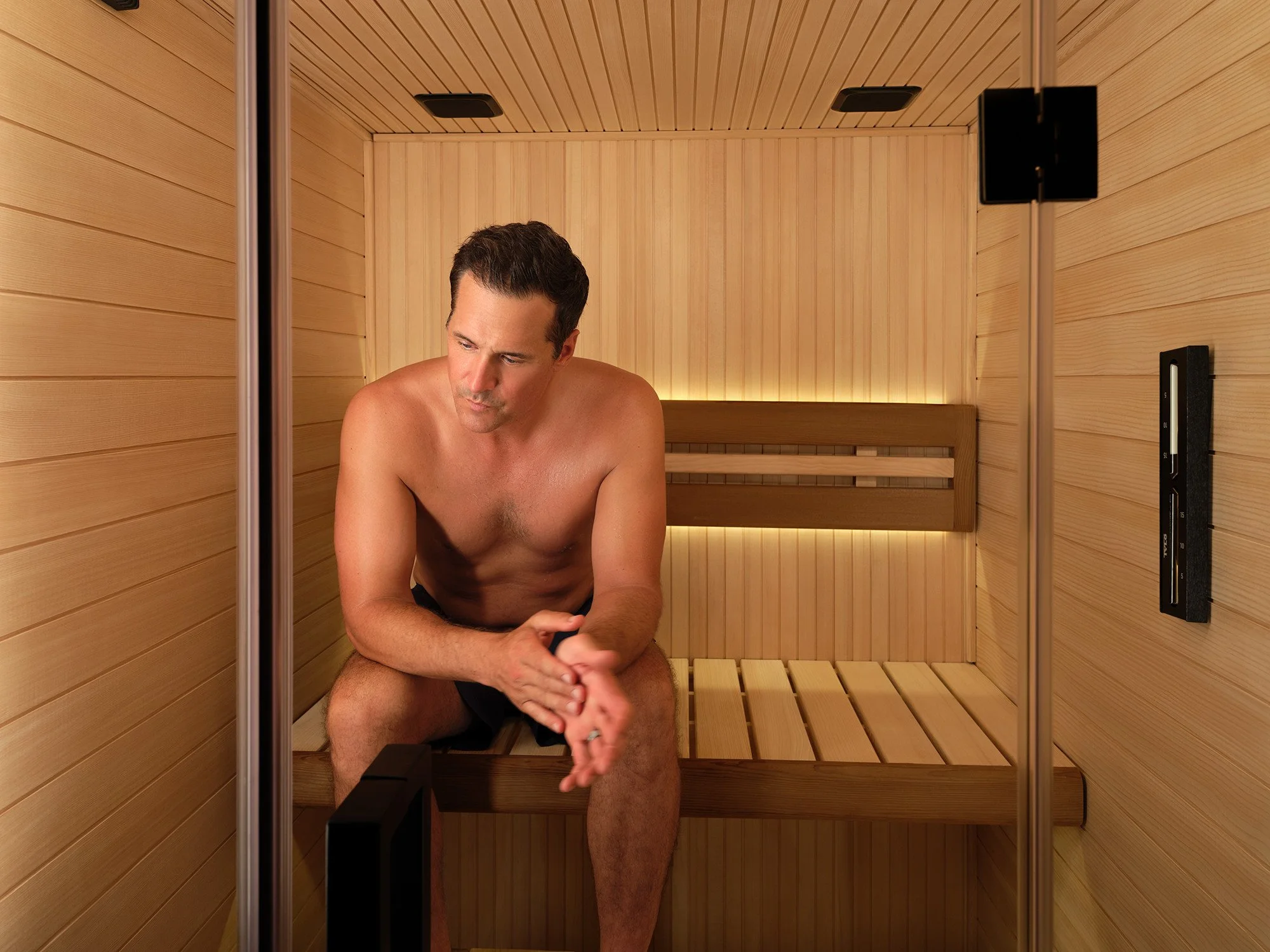 A man sitting inside a wooden sauna, shirtless, with a contemplative expression.
