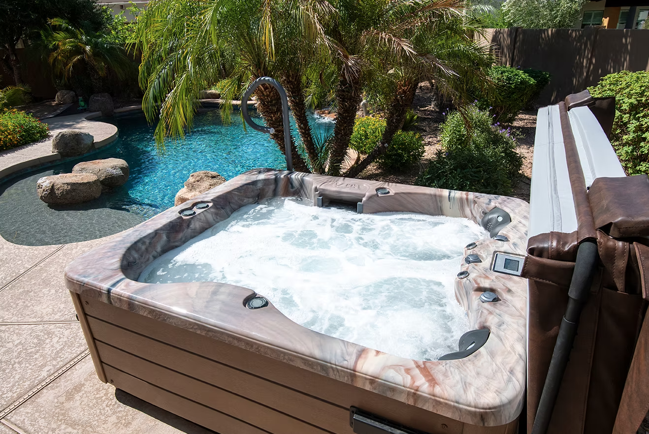 A hot tub with bubbling water beside a swimming pool with rocks and palm trees in a backyard garden.