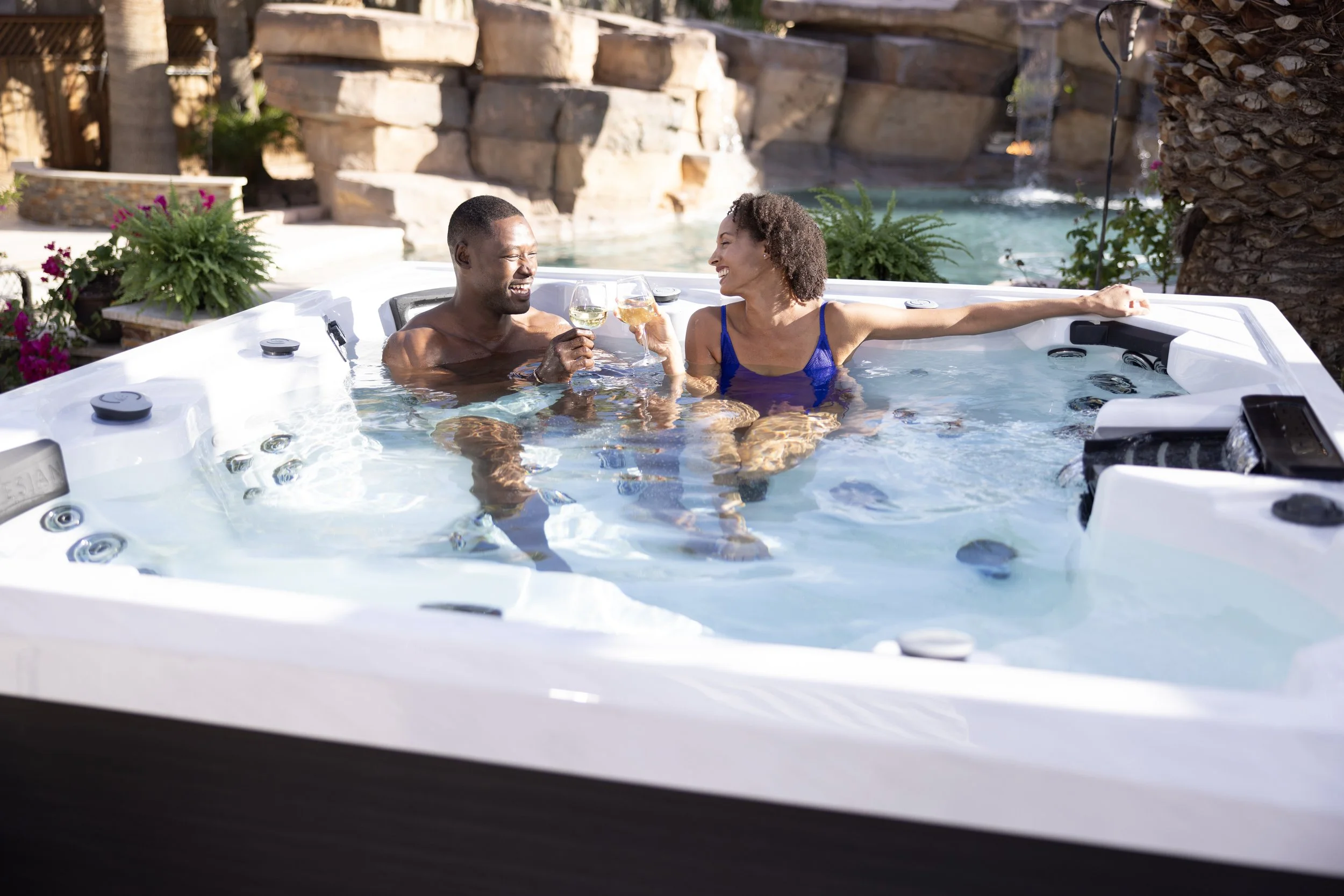 A happy couple sitting in a hot tub outdoors, clinking glasses of wine and smiling at each other, with a waterfall and lush greenery in the background.