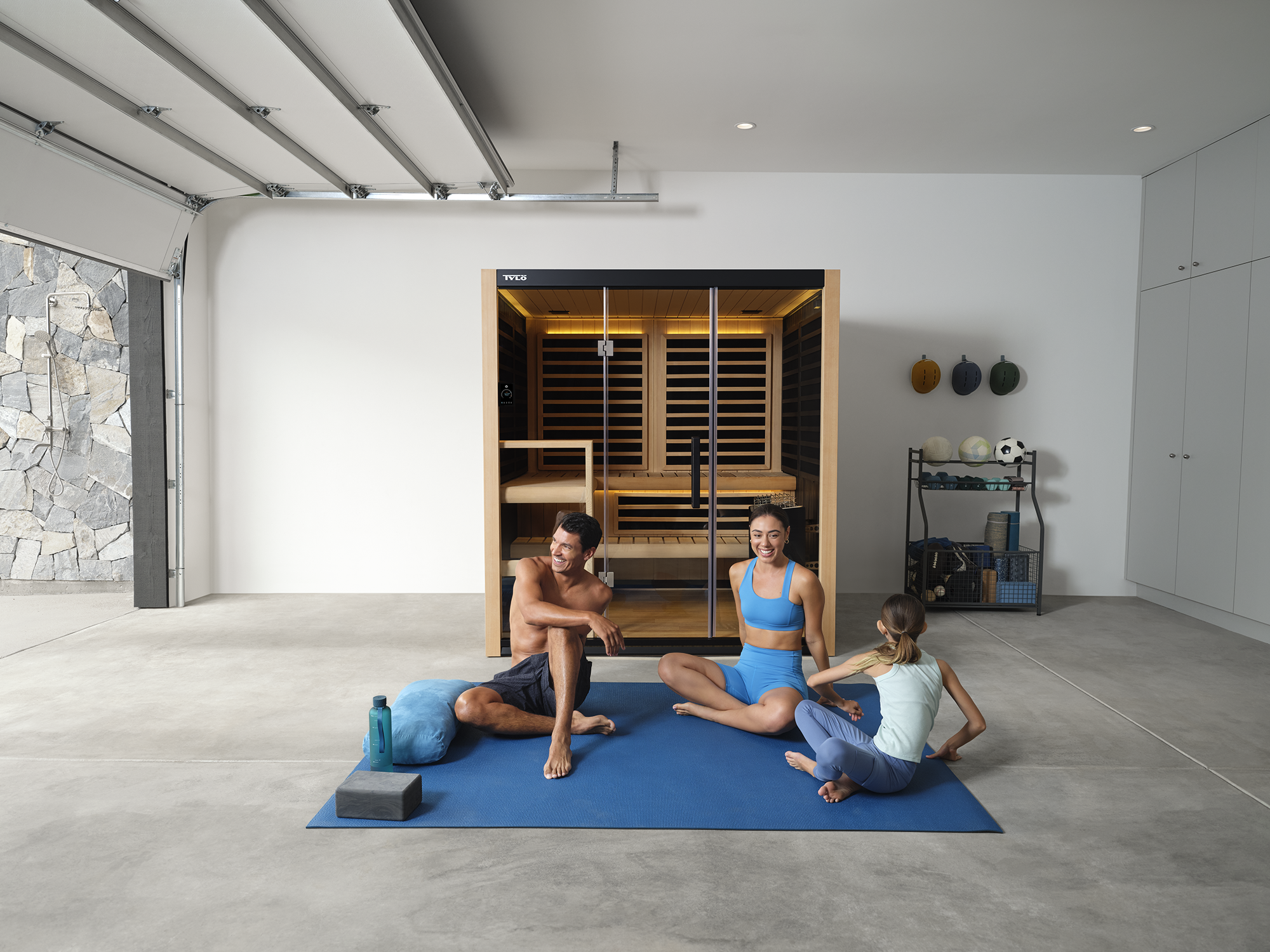A family of three, two women and one girl, sitting on a blue yoga mat in a garage with a sauna in the background. The woman in the middle is wearing a blue sports bra and leggings, the girl is in a light-colored workout top and leggings, and the man 