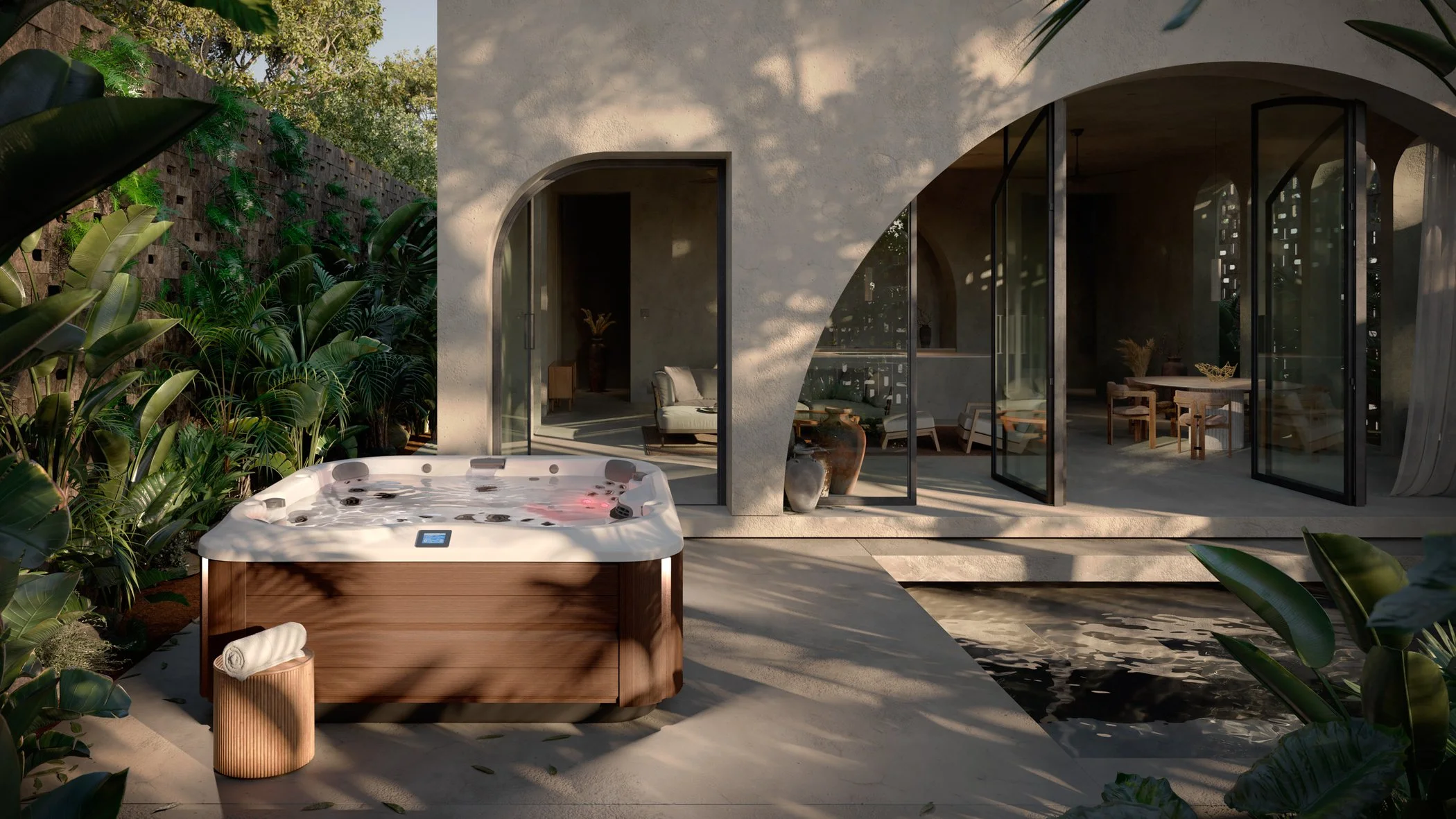 Outdoor hot tub with wooden sides, surrounded by lush tropical plants, next to a modern house with large glass doors and interior lounge and dining area visible.