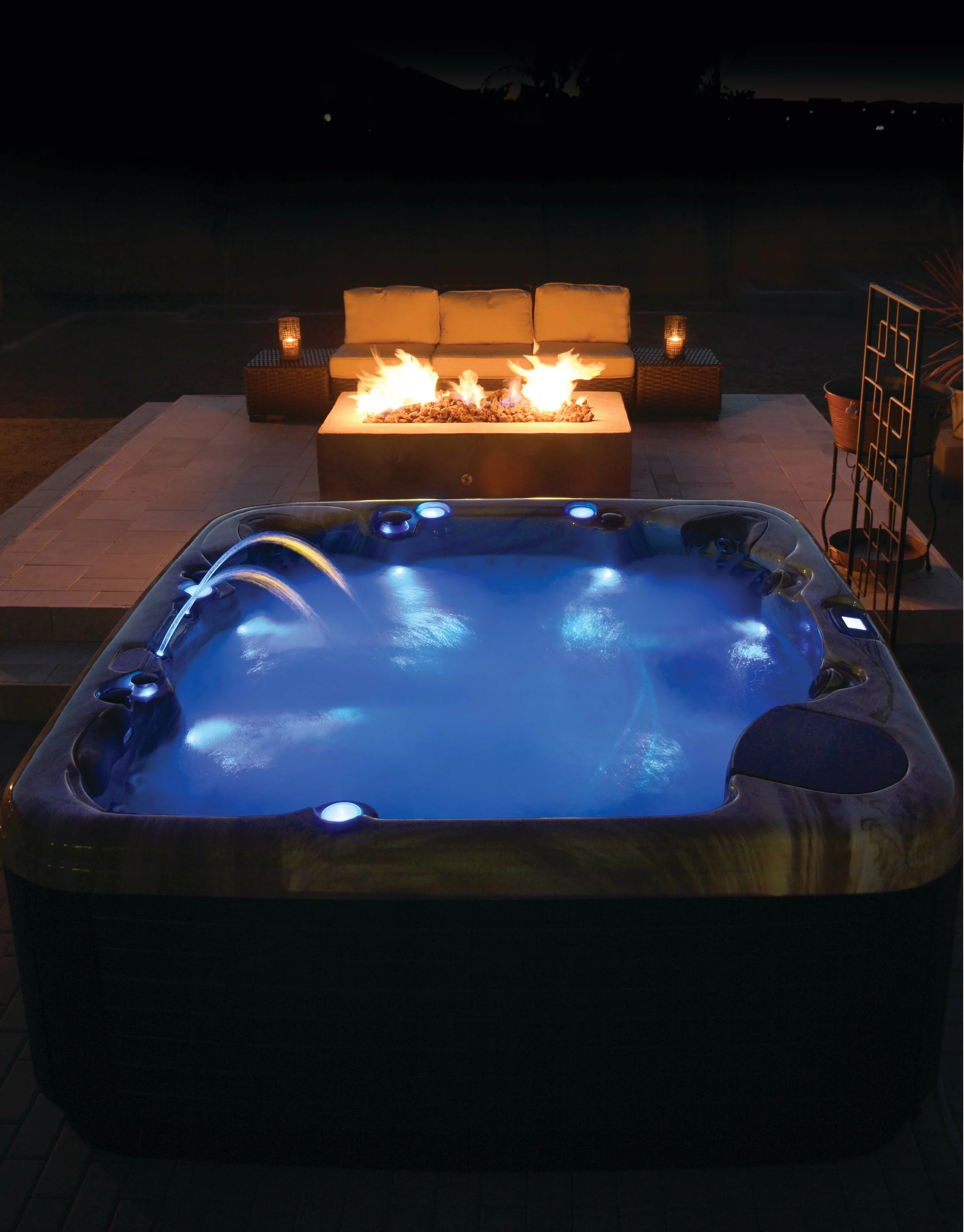 Nighttime outdoor scene featuring a lit hot tub with blue LED lights, a burning fire pit with flames, and surrounding patio furniture including a beige cushioned sofa.