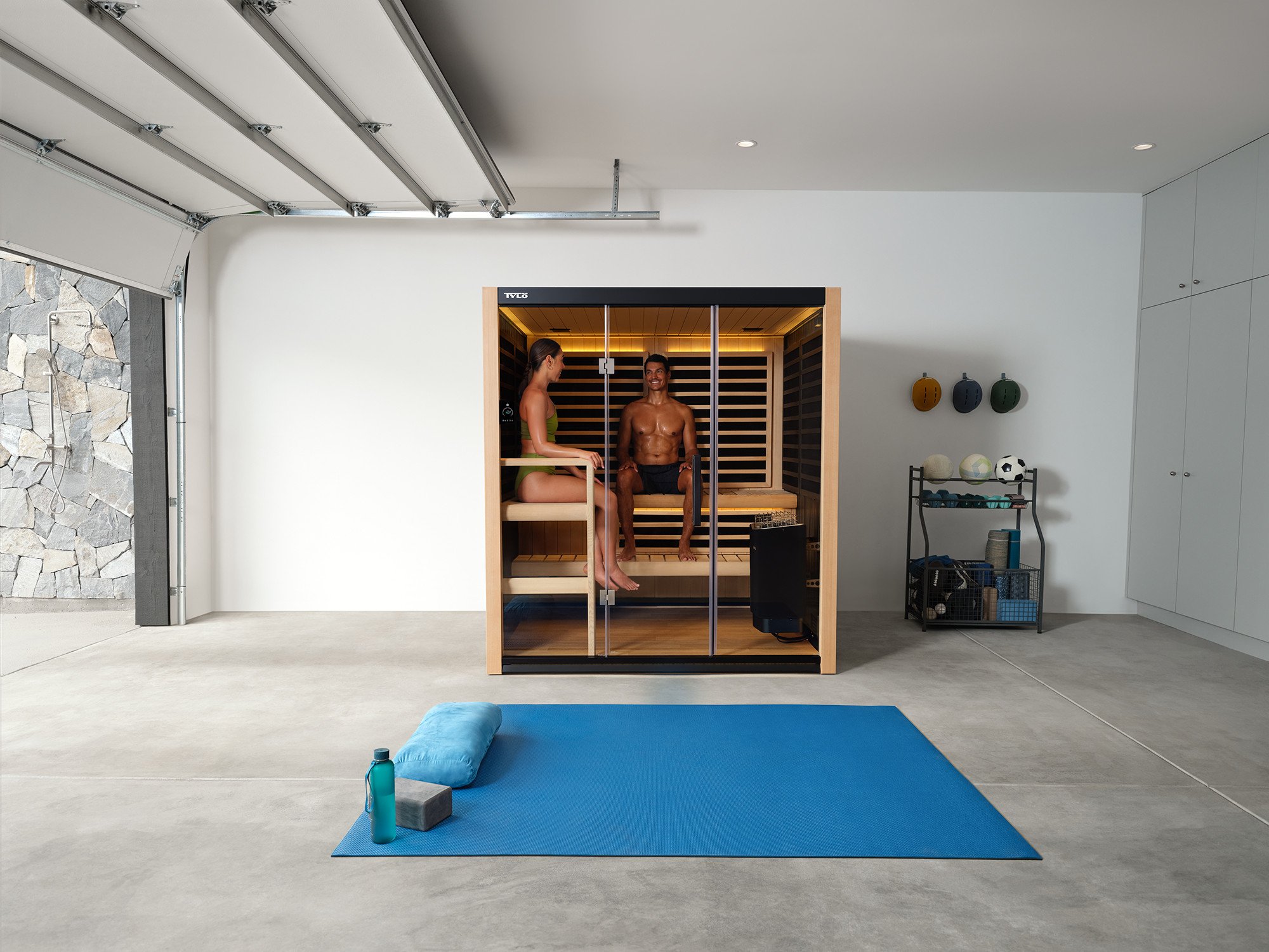 A man and woman sitting inside a wooden infrared sauna with glass doors in a modern home gym. The woman is in a green sports bra and black shorts, and the man is shirtless with black shorts. The facility has white walls, a stone wall, and fitness equ