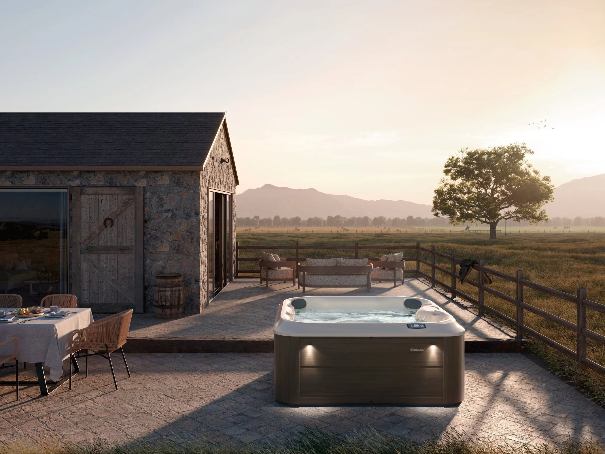 A rustic stone building with a sloped roof, overlooking a scenic countryside with mountains in the distance. The outdoor patio features a hot tub, a dining table with chairs, and a seating area with two couches. A large tree and a wooden fence are vi