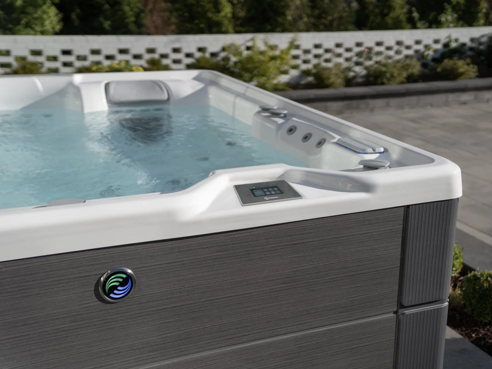 Close-up of a modern outdoor hot tub with digital controls on a patio, with a garden and white lattice fence in the background.