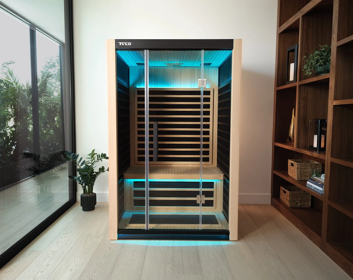 A modern infrared sauna with glass doors in a well-lit room near a large window, wooden flooring, and bookshelf with decorative items.