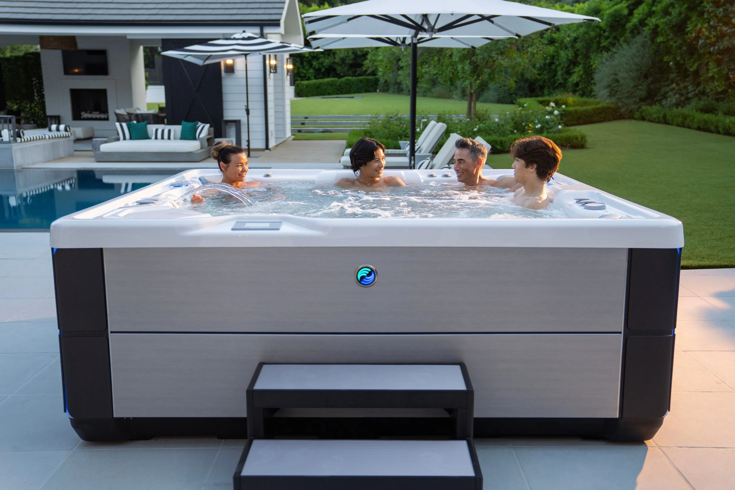 A group of five people enjoying a hot tub in a backyard with a swimming pool, patio furniture, umbrellas, and lush green landscaping.