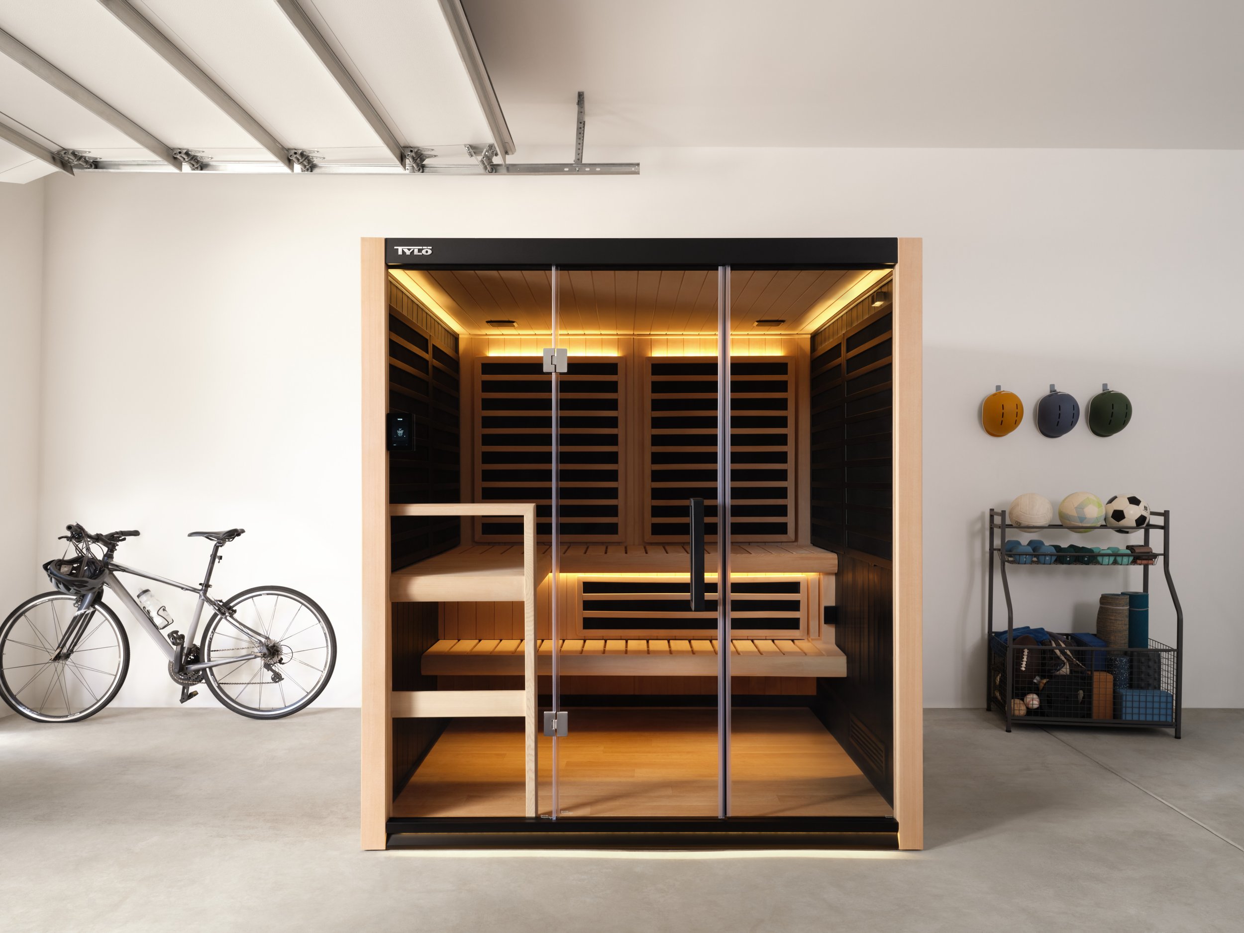 Modern home sauna with glass door, wooden interior, and black exterior, in a minimalist garage or fitness room with a bicycle and exercise equipment.