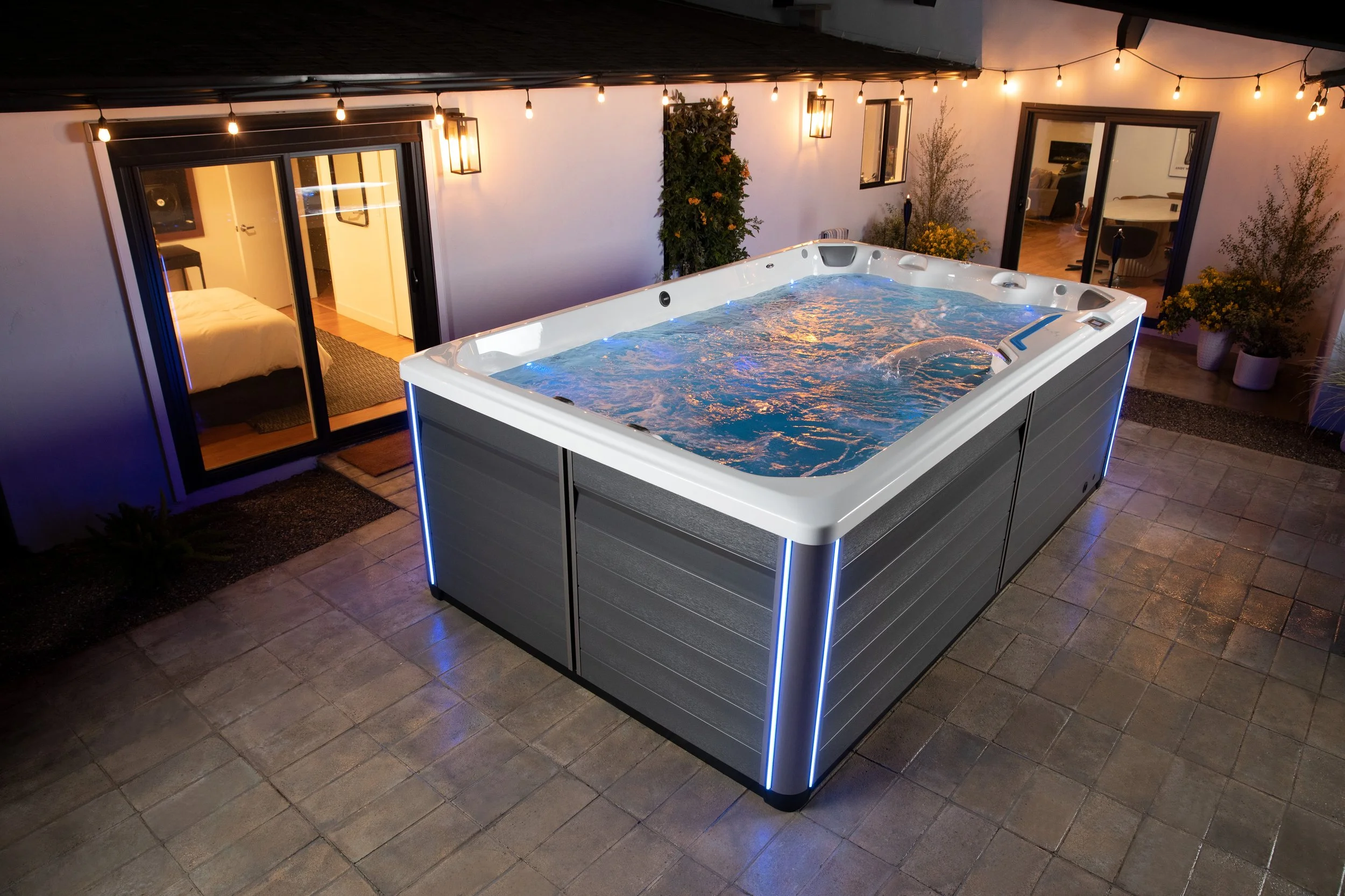 An outdoor patio with a swim spa illuminated by blue LED lights, string lights hanging from above, potted plants, and sliding glass doors leading to indoor rooms.