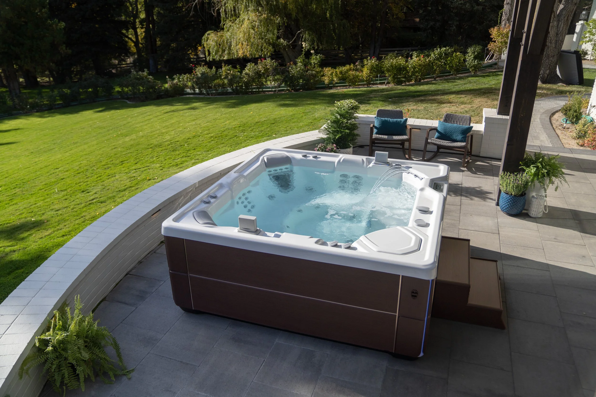 Outdoor hot tub on patio with seating area, surrounded by a green lawn and trees.