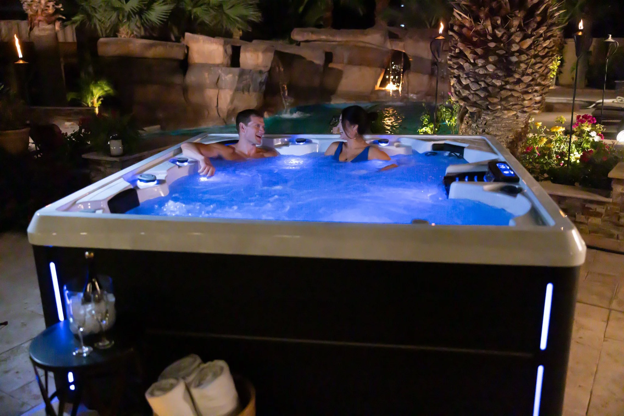 Couple enjoying a relaxing moment in a lit-up hot tub at night outdoors, surrounded by tropical plants and decorative lighting.