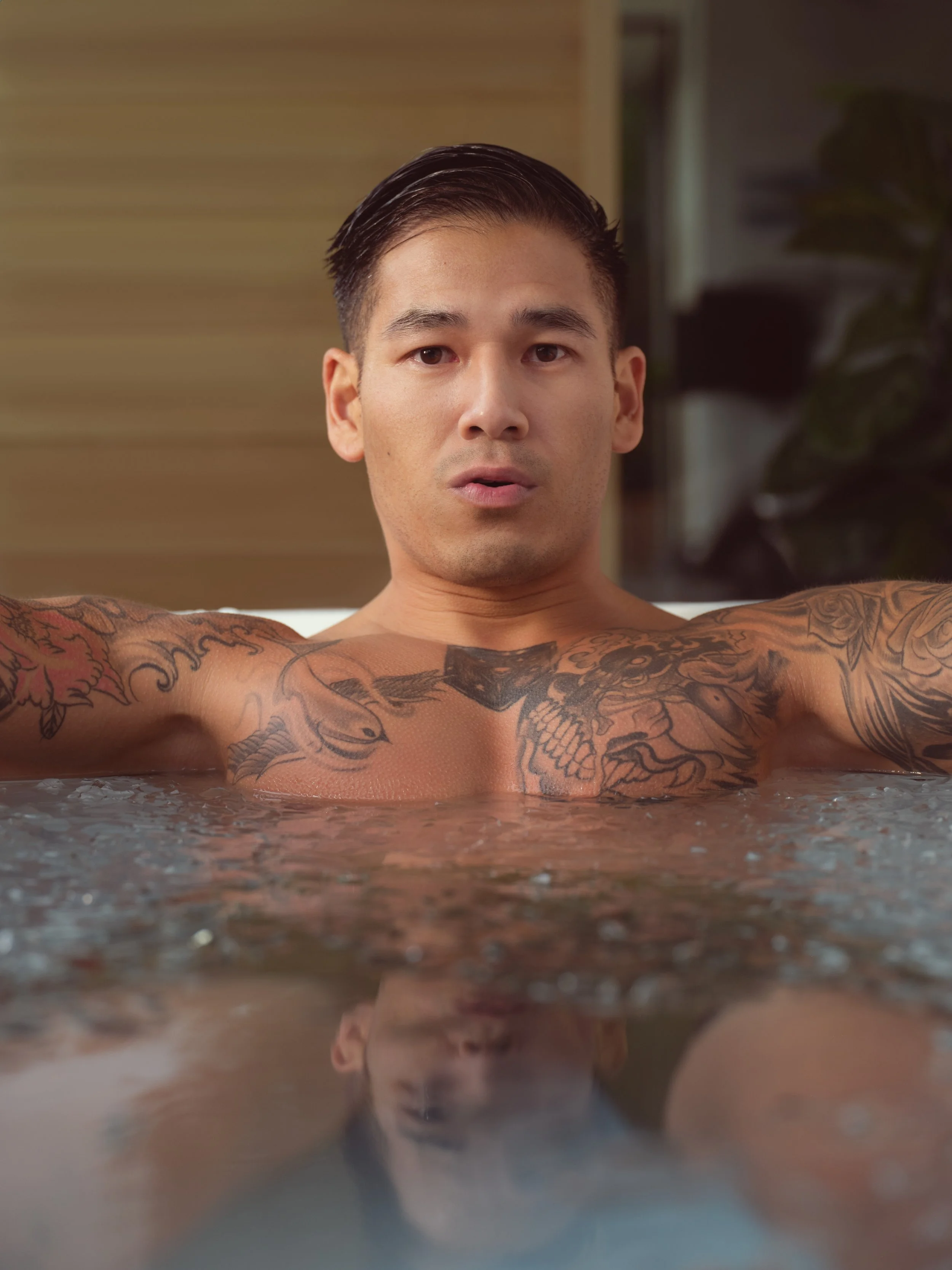 A shirtless man with tattoos on his chest and arms, submerged in a cold plunge, looking directly at the camera with a determined expression.