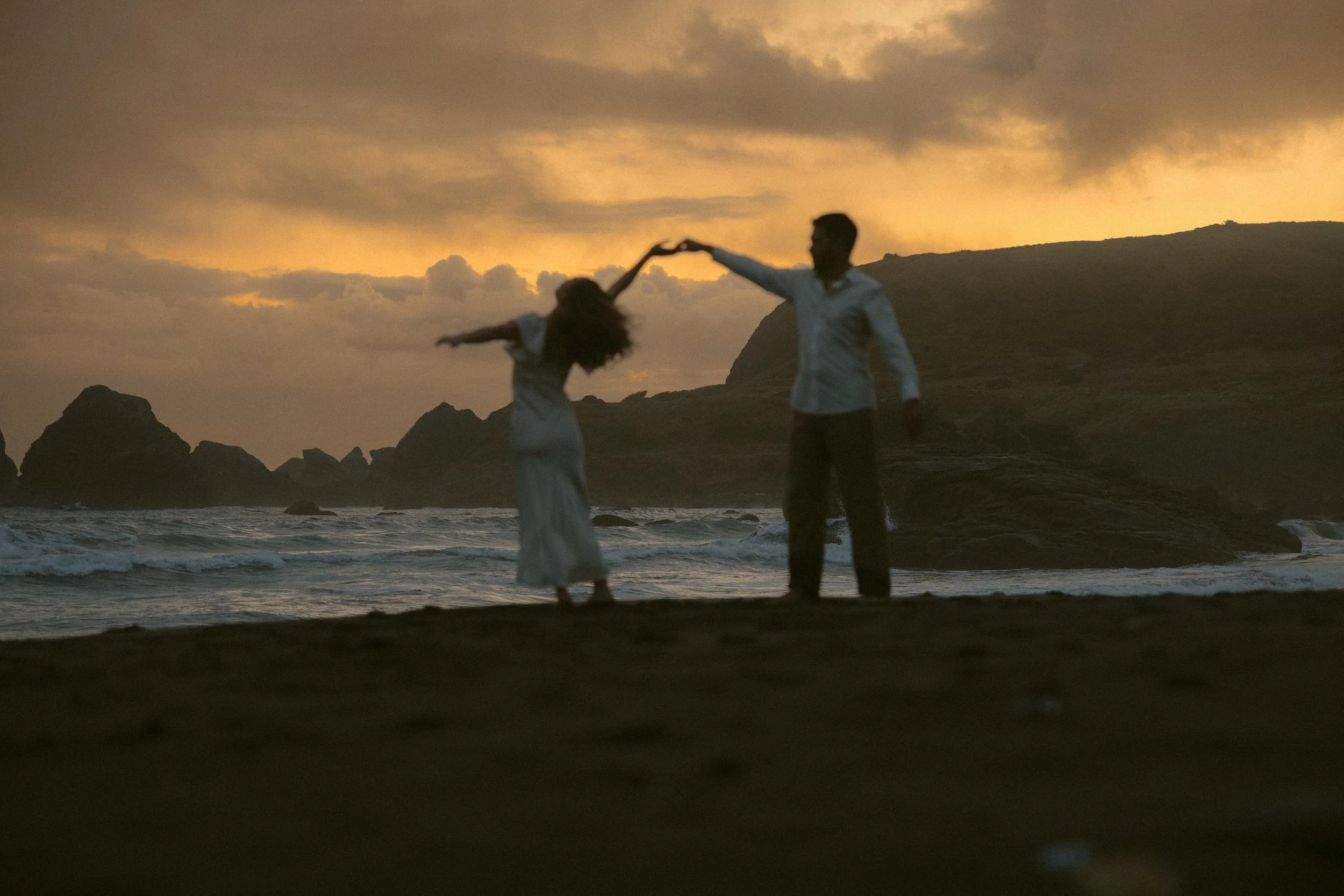 A couple dancing on a beach at sunset with a rocky landscape in the background.