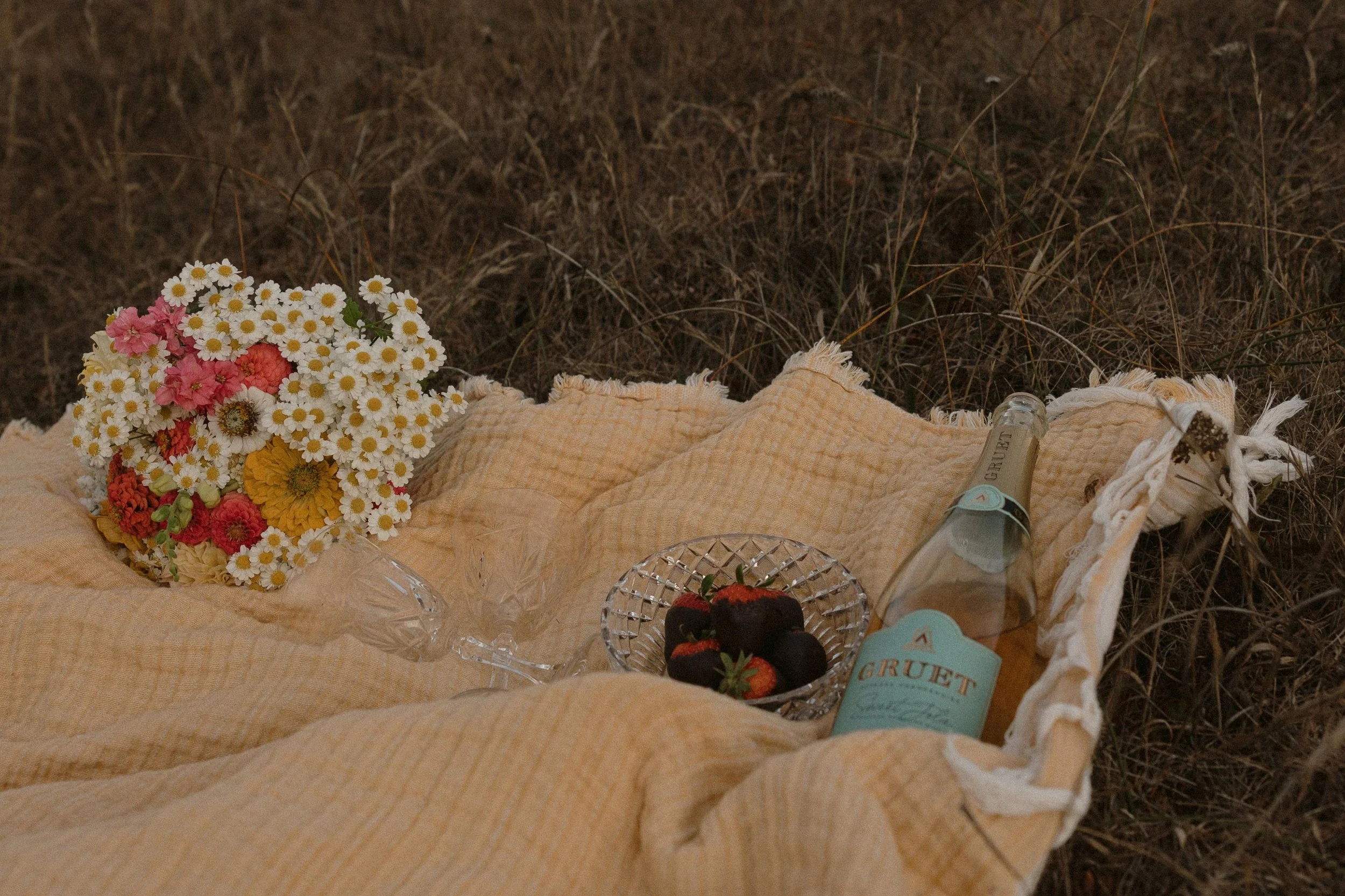 A picnic setup on a beige blanket with a bouquet of mixed flowers, strawberries in a glass bowl, and a couple of bottles with blue labels, inside a grassy outdoor area.