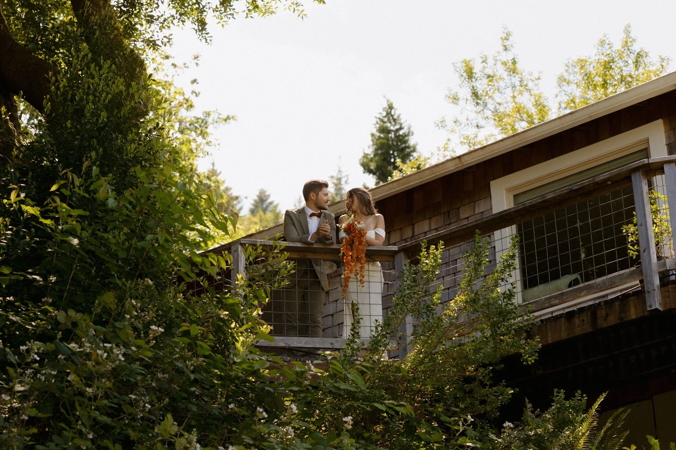 A couple dressed in wedding attire standing on a balcony, surrounded by lush greenery and trees, with sunlight filtering through the leaves.