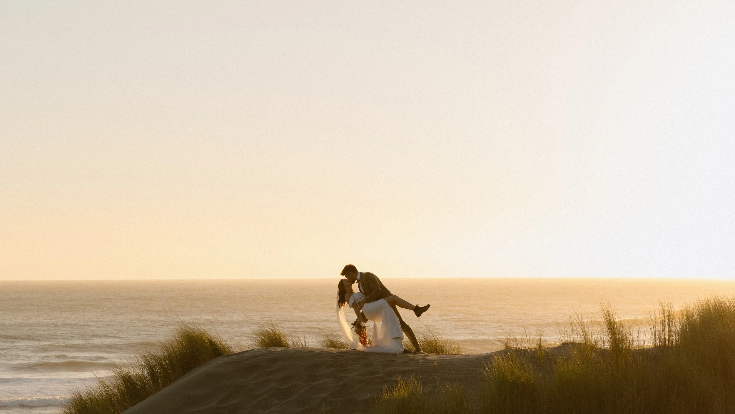 A couple kissing on the beach during sunset, with the man holding the woman in his arms and both in wedding attire.