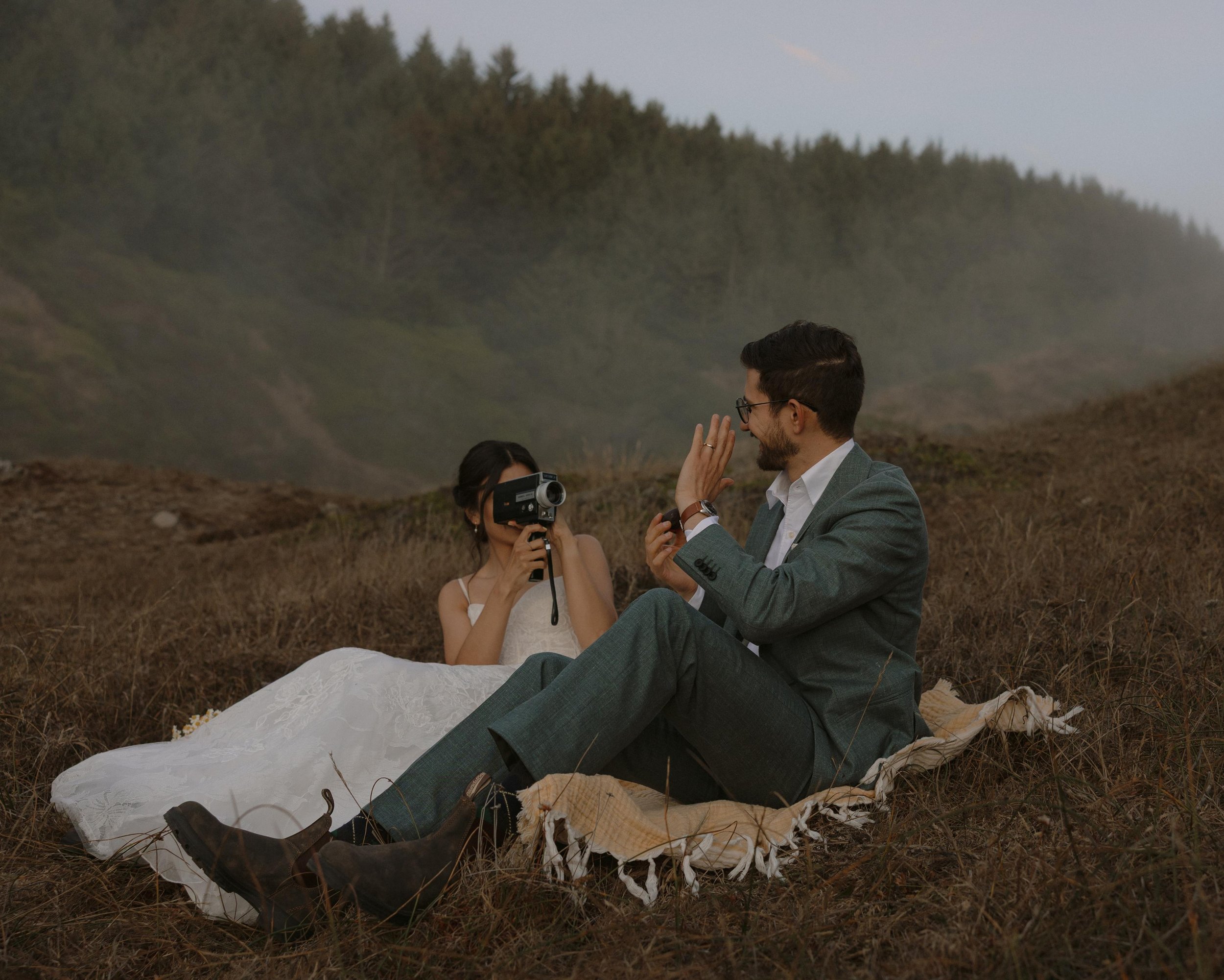 A couple sitting on the ground outdoors at sunset, with the man wearing a suit and the woman in a white dress, possibly during a wedding or engagement photoshoot, with trees and a cloudy sky in the background.