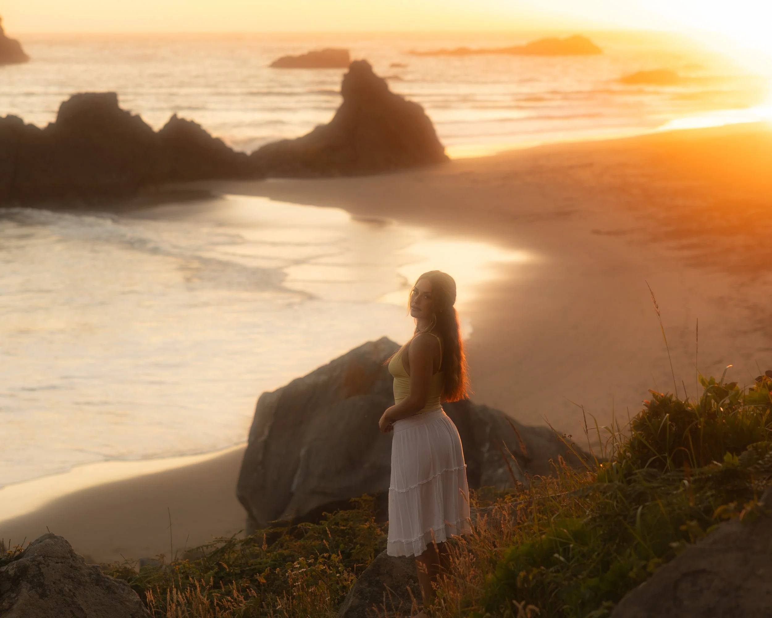 A woman with long hair standing on a rocky beach during sunset, wearing a white dress, with the ocean and rocks in the background.