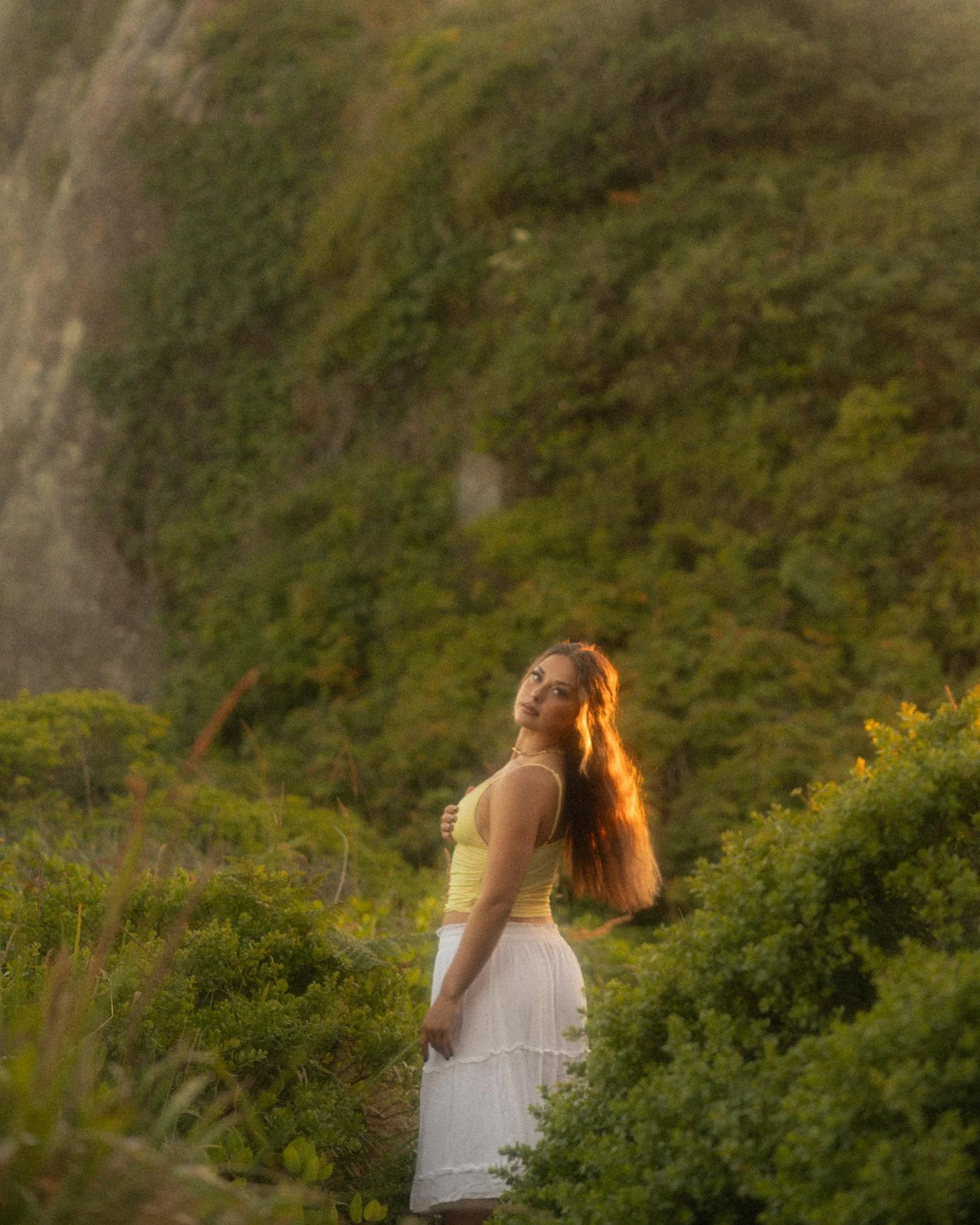 A woman with long, wavy hair wearing a yellow top and a white skirt standing outdoors at sunset, surrounded by green bushes and trees.