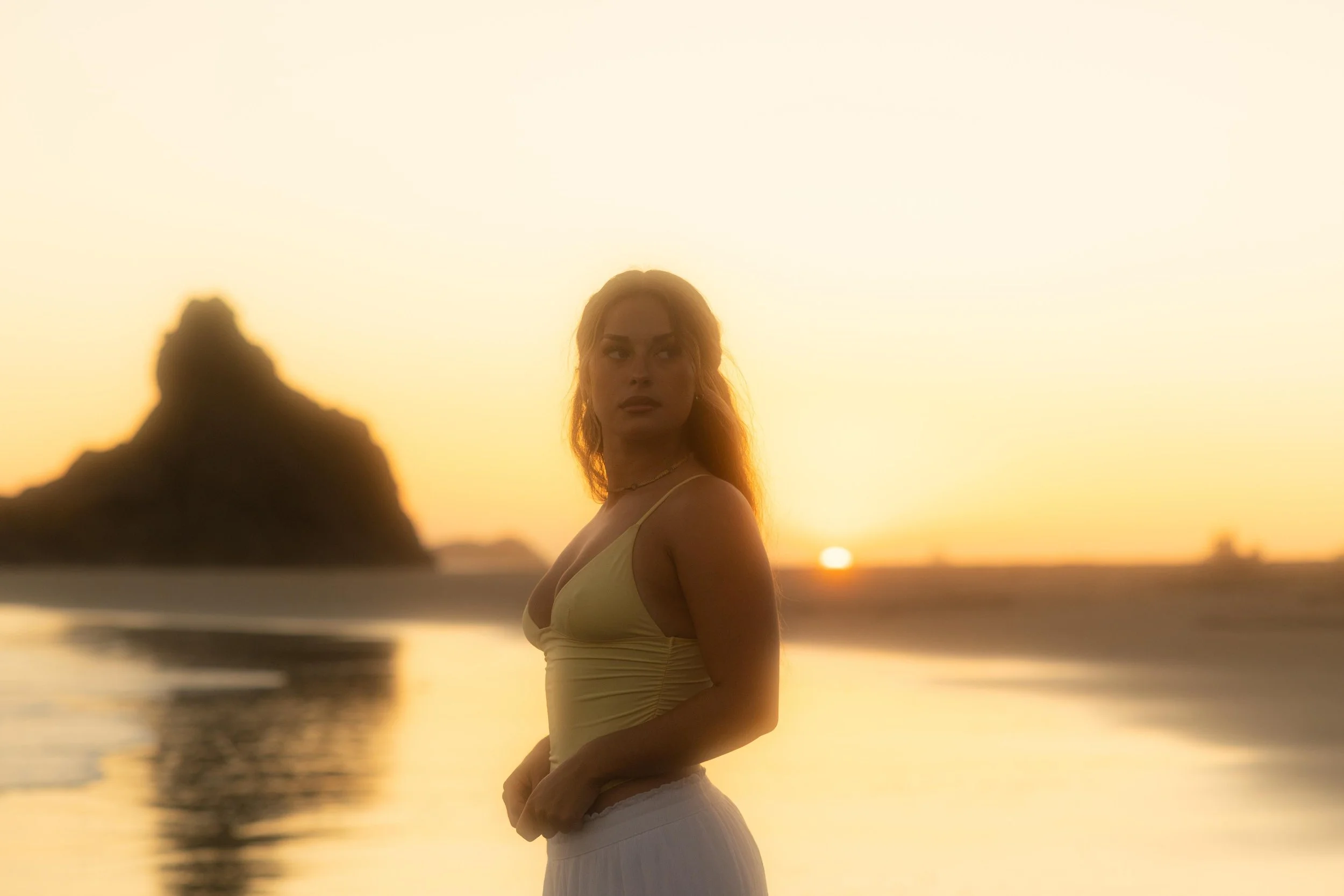 A woman with blonde hair in a tank top and skirt standing on the beach during sunset, with a rocky formation in the background and the sun near the horizon.