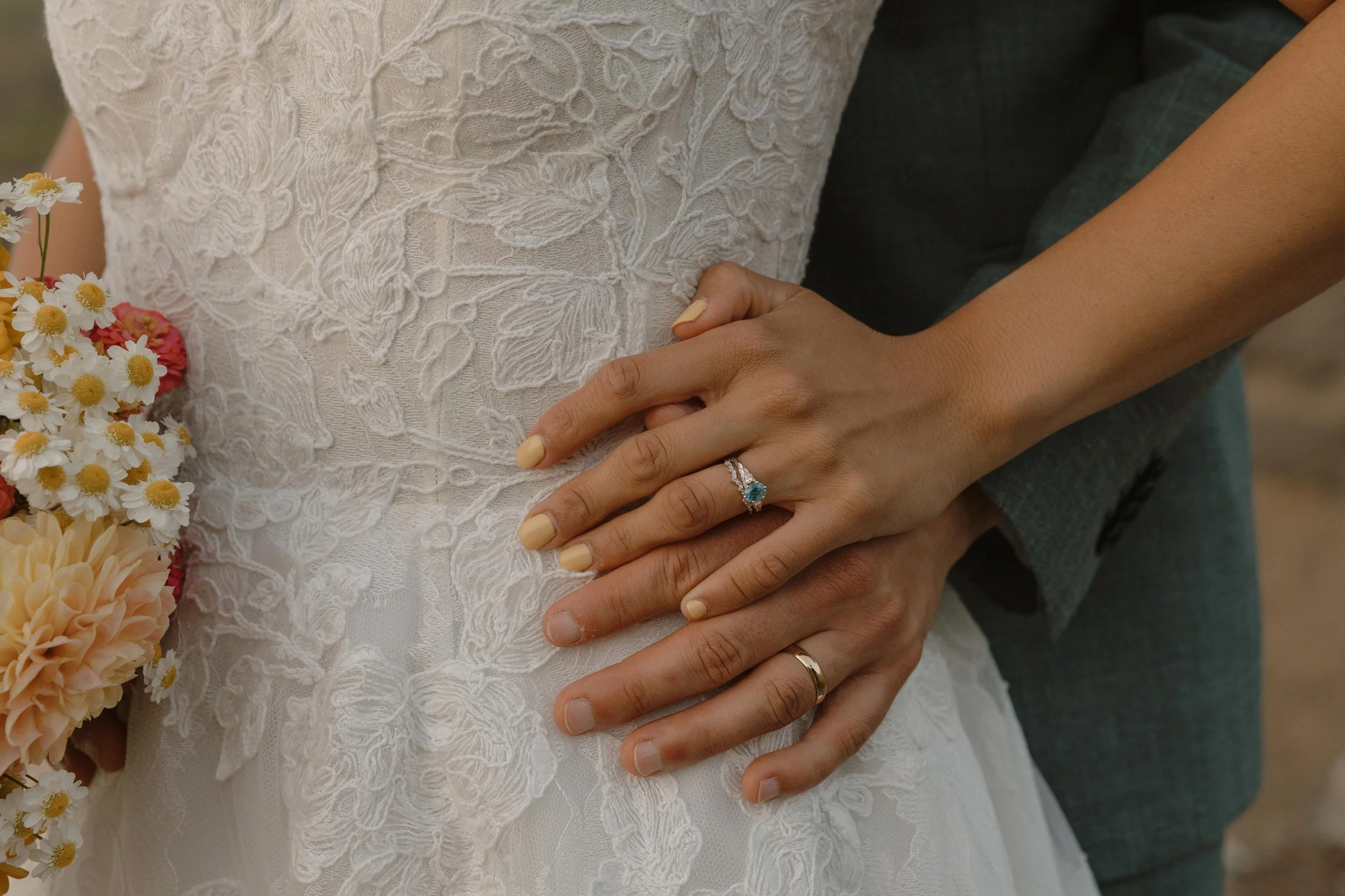 Close-up of couple's hands with wedding rings resting on a bride's lace wedding dress, with a bouquet of white and pink flowers nearby.