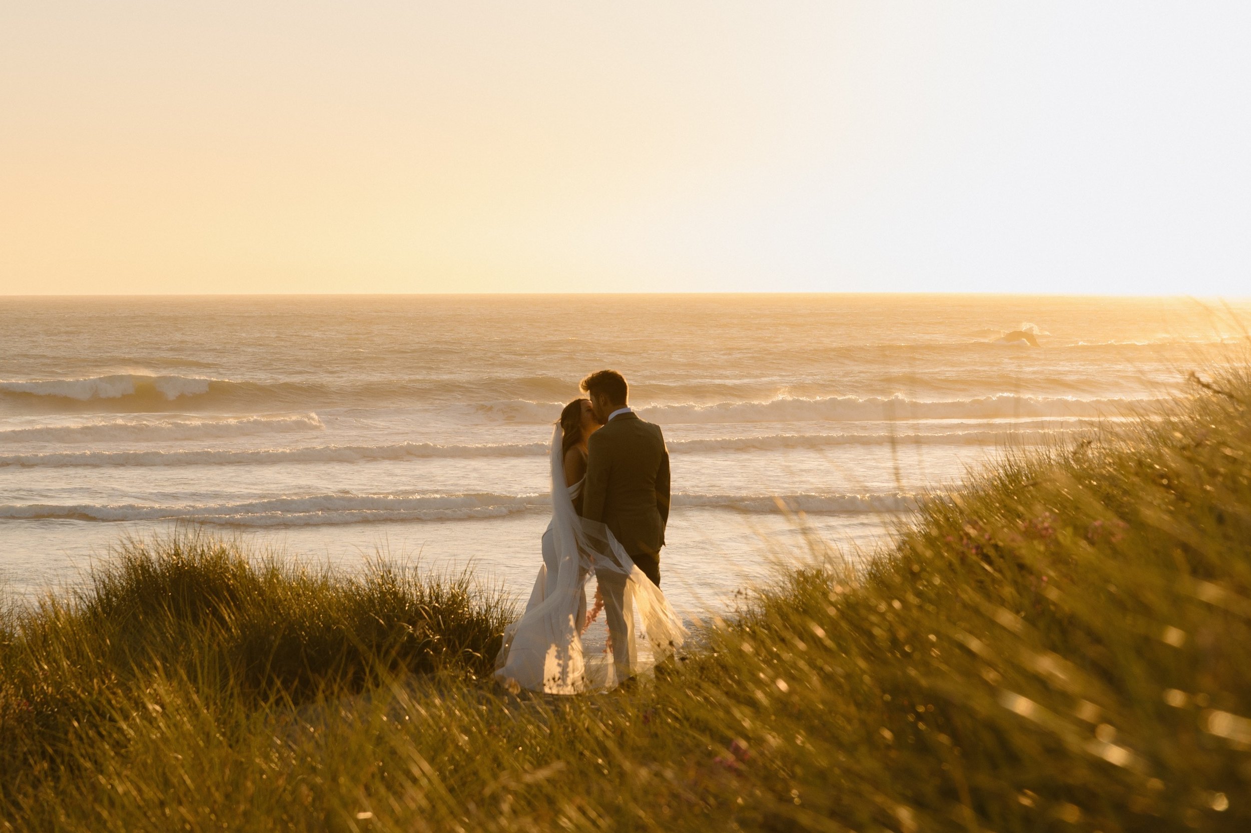 A bride and groom standing closely together on a beach during sunset, embracing and kissing with the ocean waves in the background.