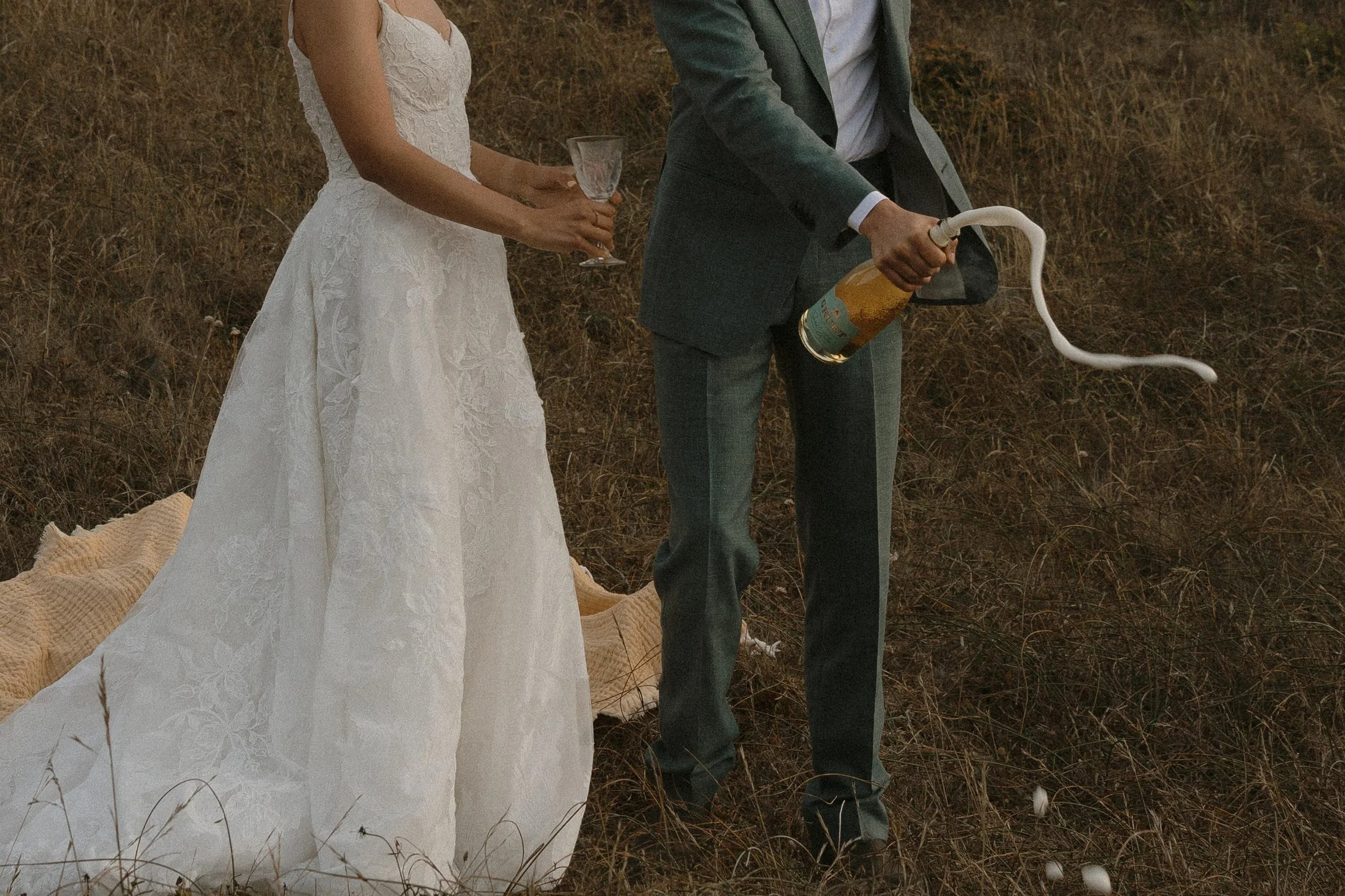 A bride in a white wedding dress holding a glass of champagne, standing next to a groom in a gray suit holding a banana-shaped balloon with a wavy white string attached, outdoors on a dry grass field.