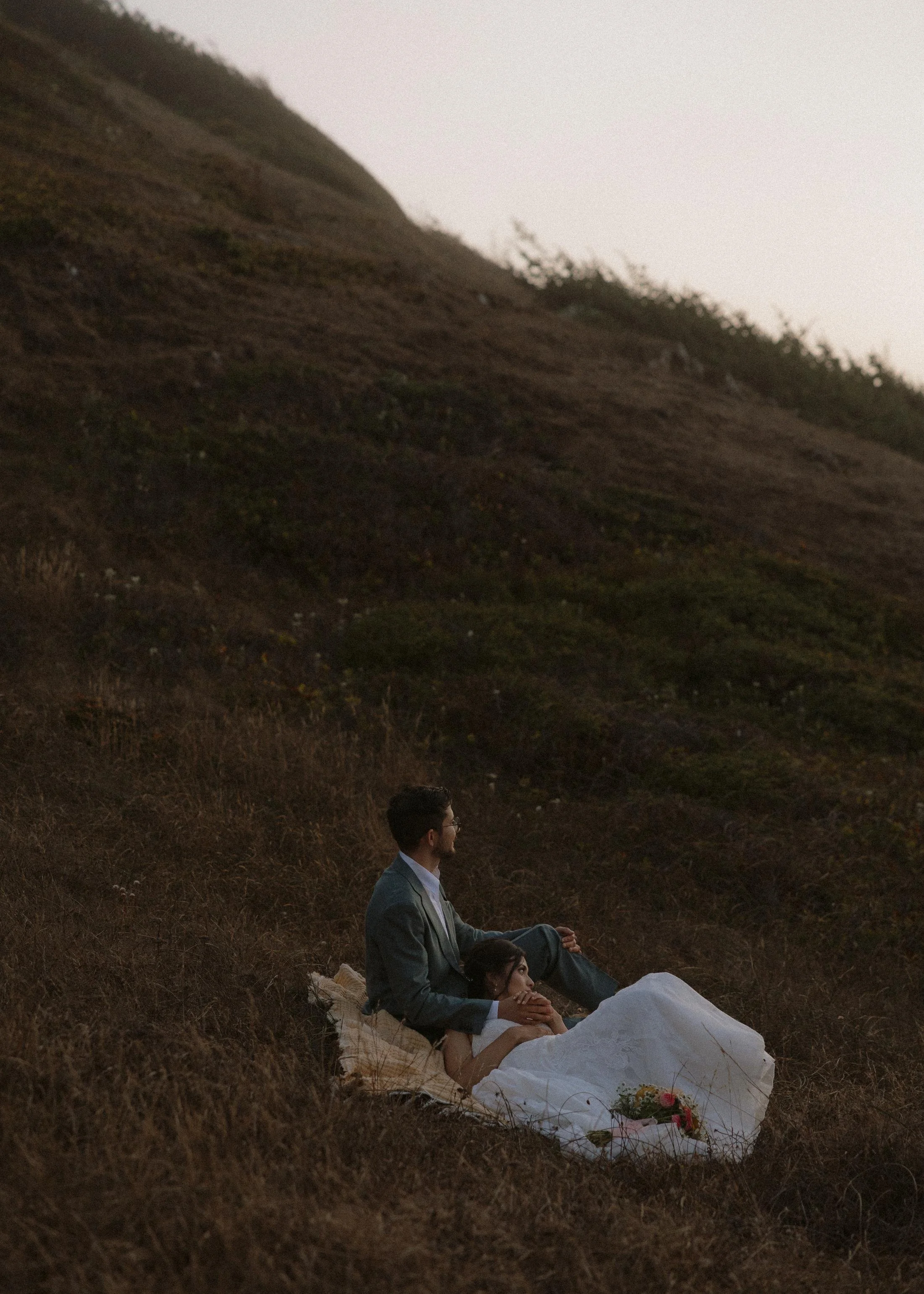 A man in a suit and glasses and a woman in a white dress sitting on a grassy hillside during dusk, with a large hill or mountain in the background.