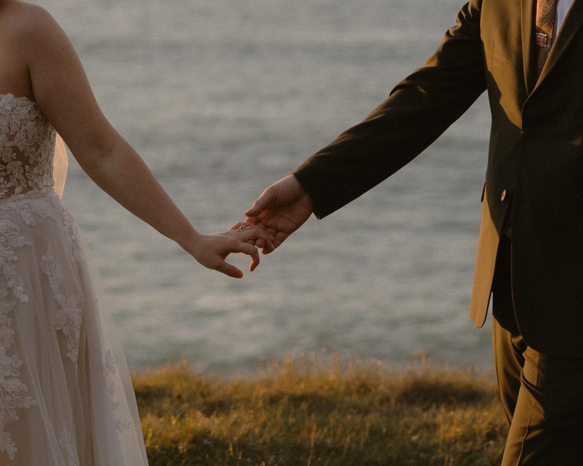Close-up of a bride and groom holding hands over a body of water, with the bride in a white lace wedding dress and the groom in a brown suit and yellow pants, during sunset.