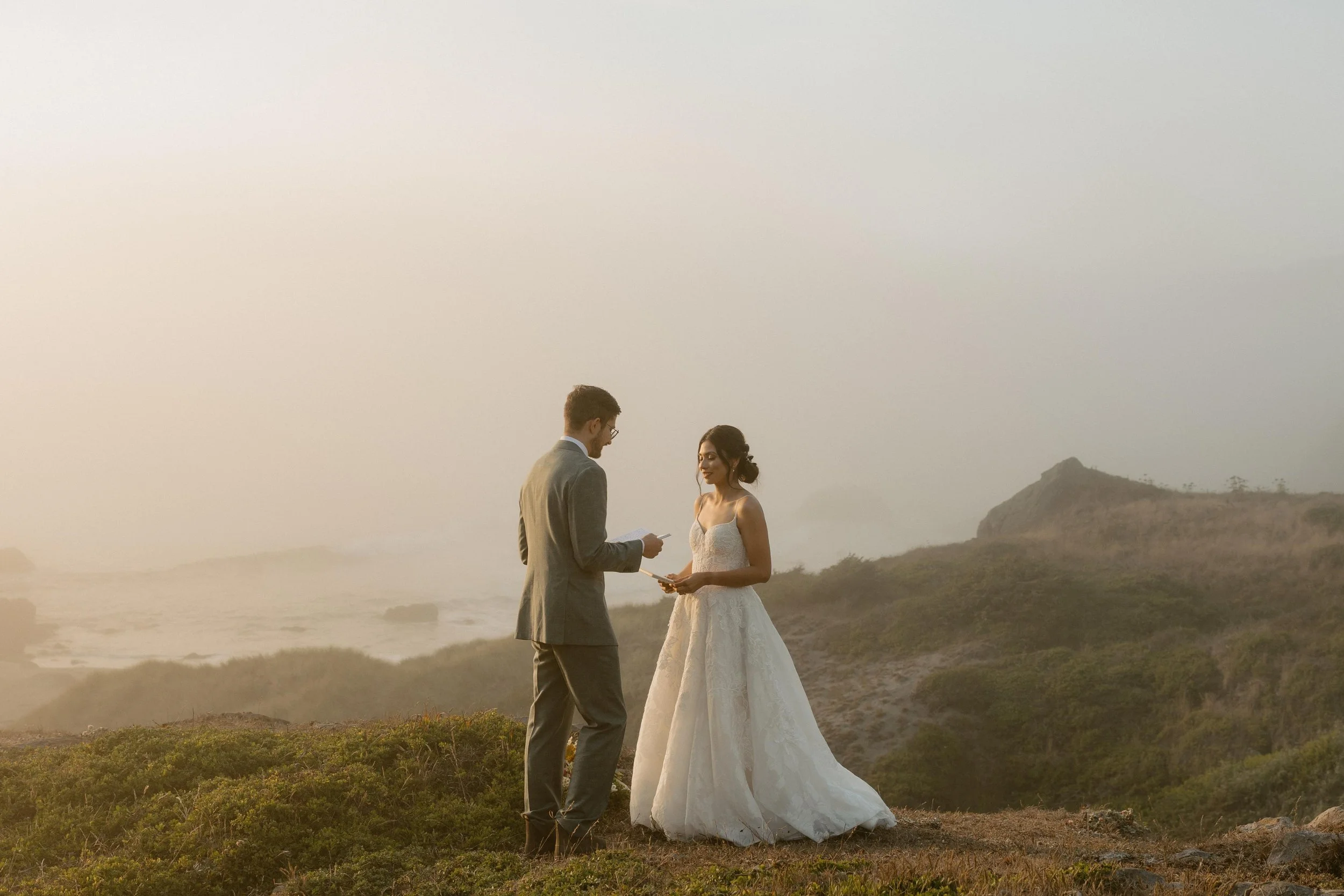 A couple exchanging vows outdoors during sunset, with scenic hills in the background.